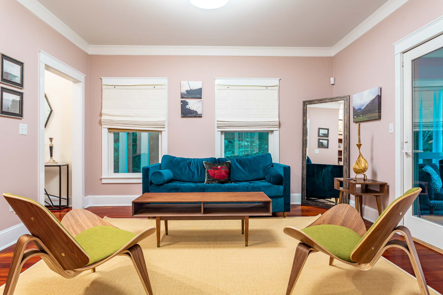 A modern living room with light pink walls, a teal sofa, a wooden coffee table, and two unique wooden chairs with green cushions on a yellow rug, featuring large windows with roman shades.