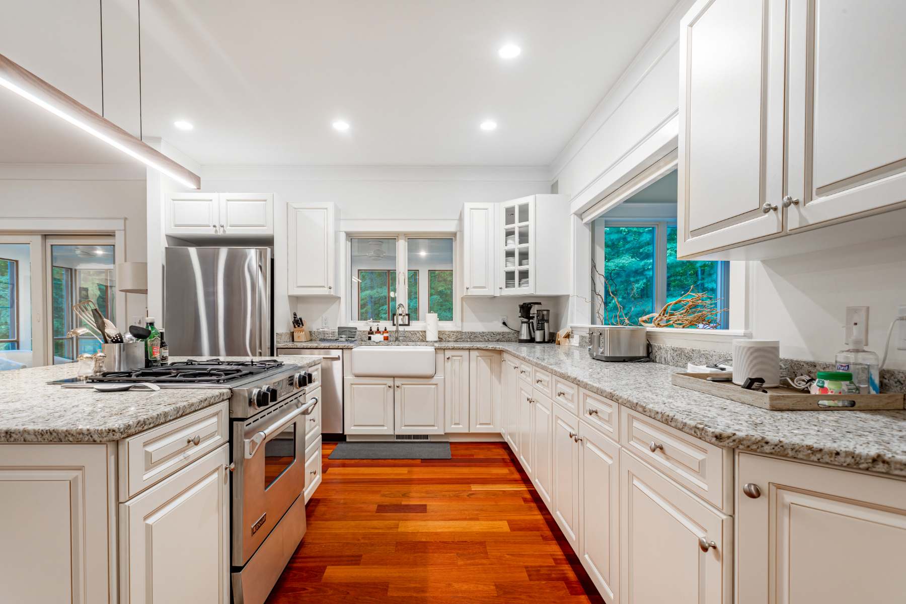 Spacious white kitchen featuring extensive granite countertops, white shaker cabinets, a farmhouse sink under a window, and stainless steel appliances, with warm hardwood floors throughout.