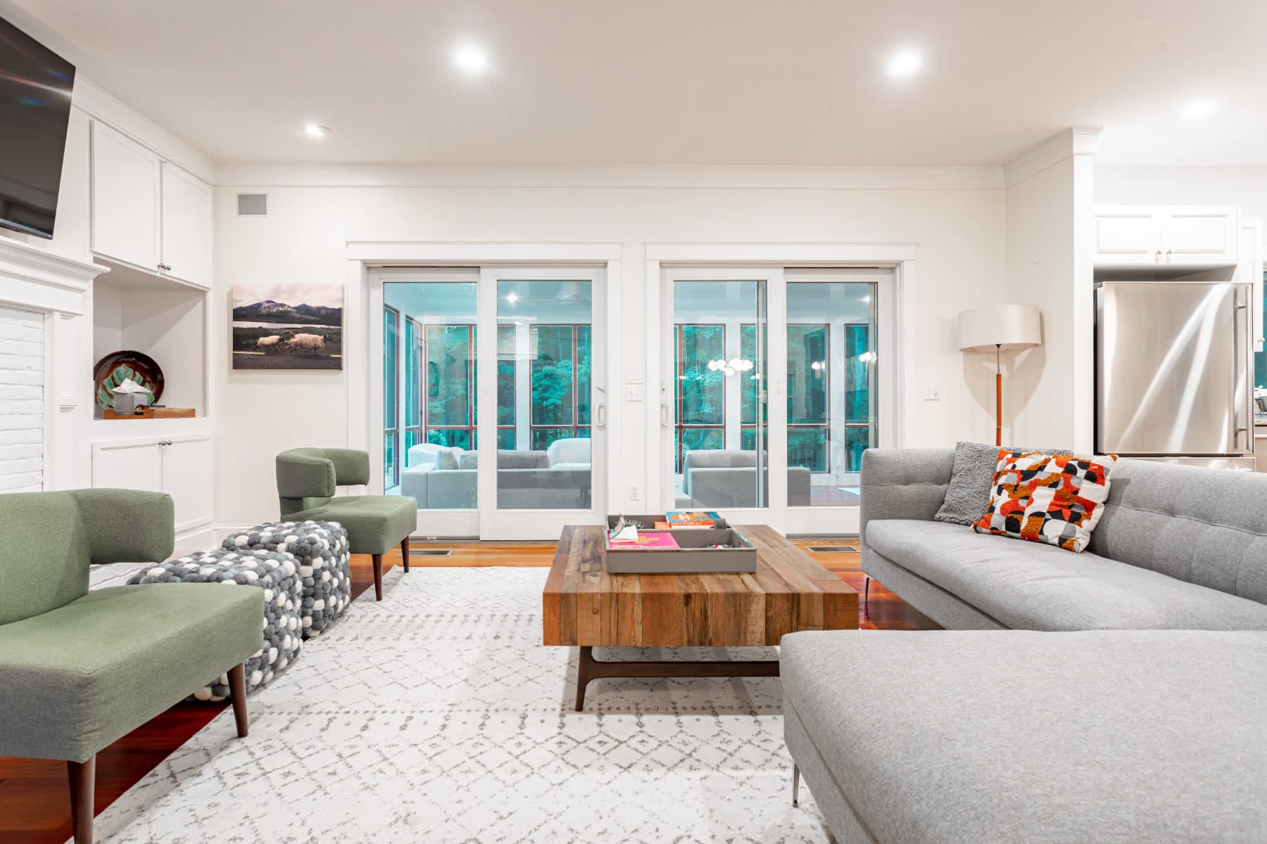 Modern living room with a gray sectional sofa and a wooden coffee table on a light patterned rug, facing a white brick fireplace and built-in shelves, with sliding glass doors leading to another room.