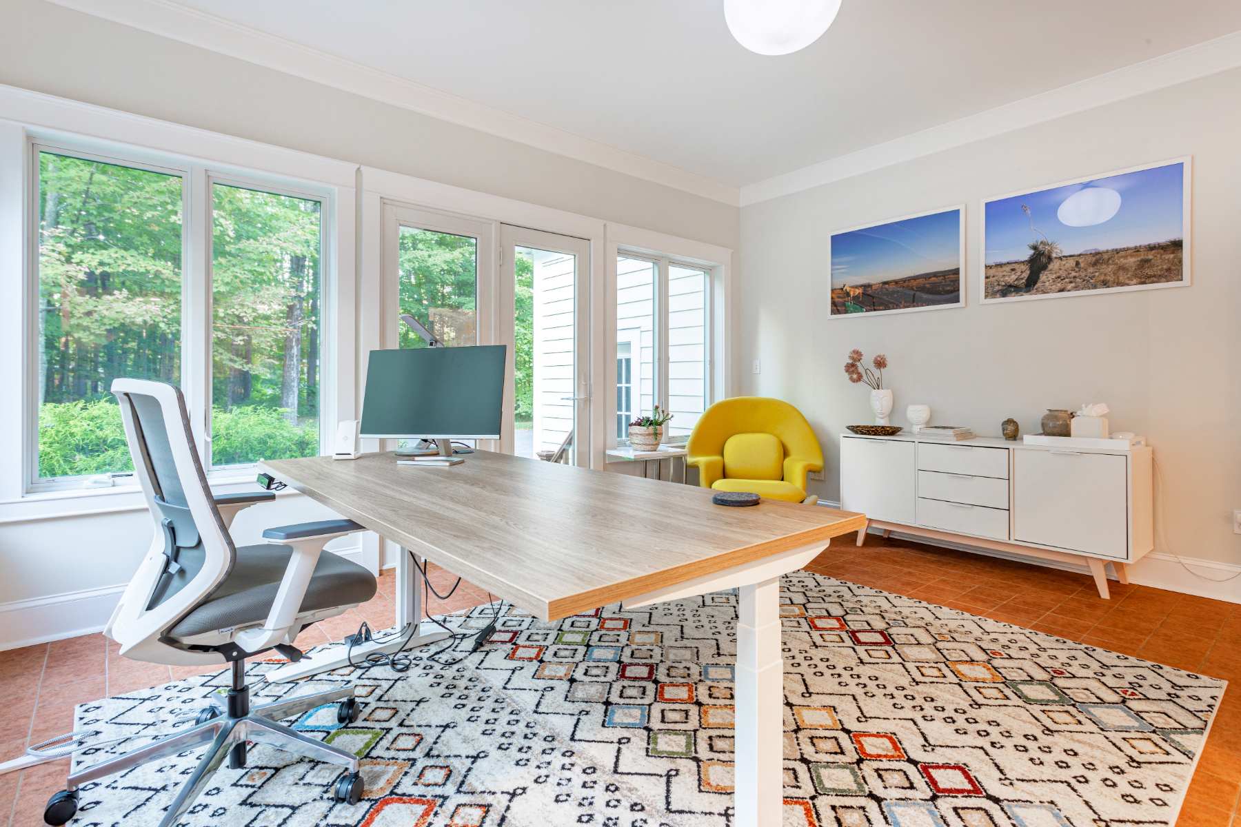 A bright home office with large windows offering views of the outdoors, featuring light-colored walls and a colorful geometric rug over tiled flooring. A modern standing desk with an ergonomic chair and a yellow accent armchair complete the space.