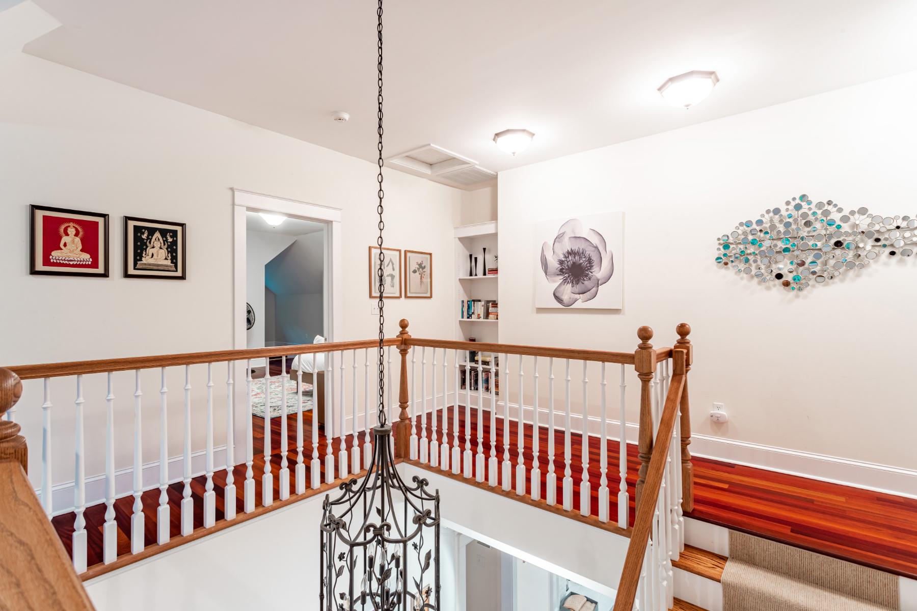 An upstairs landing area with polished hardwood floors, a white and wood railing overlooking a lower level, and various artworks on the white walls, illuminated by a decorative chandelier.