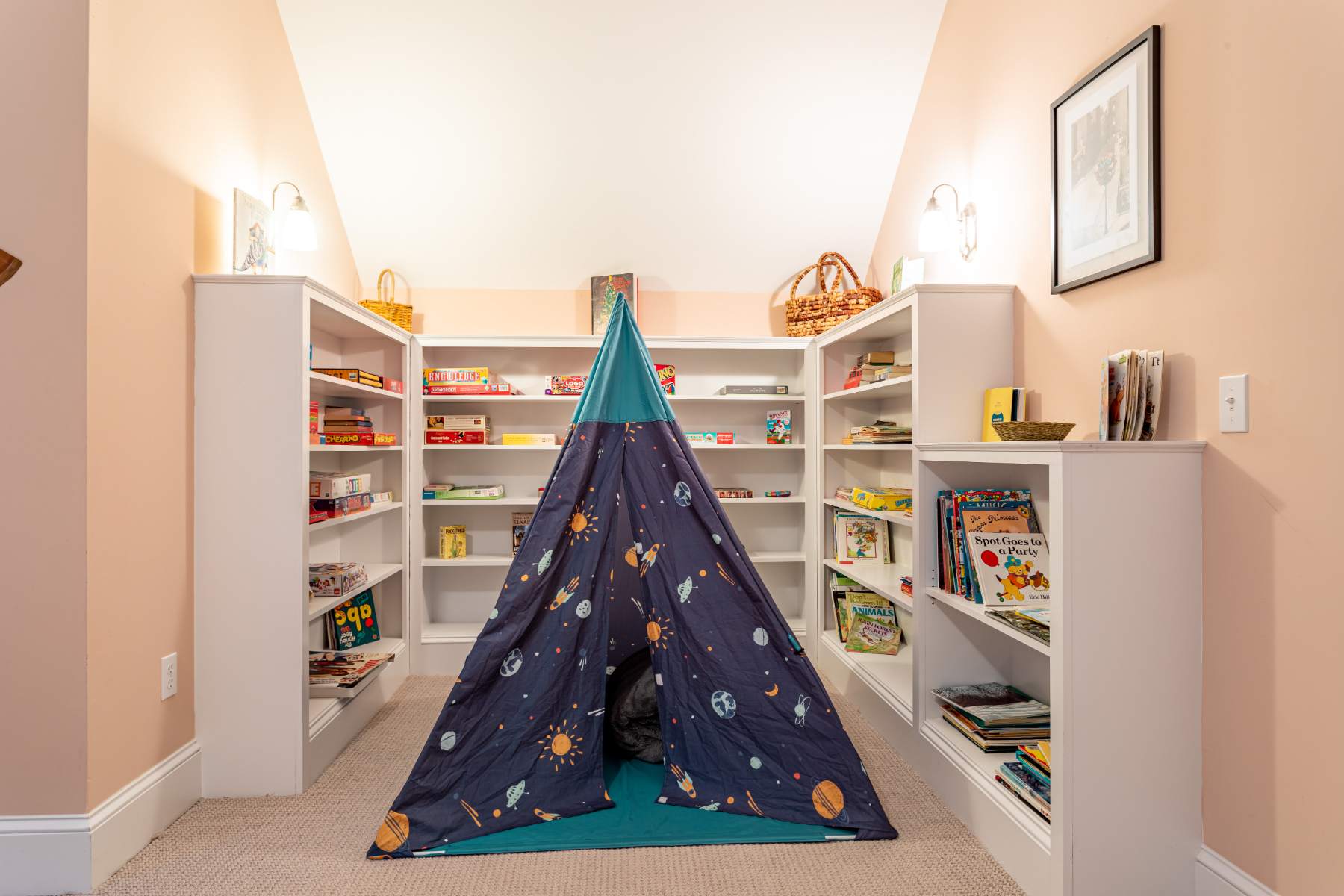 A dedicated kids' play area featuring a blue and green tent surrounded by white built-in bookshelves filled with books and games. The room has sloped ceilings and warm lighting from wall sconces.