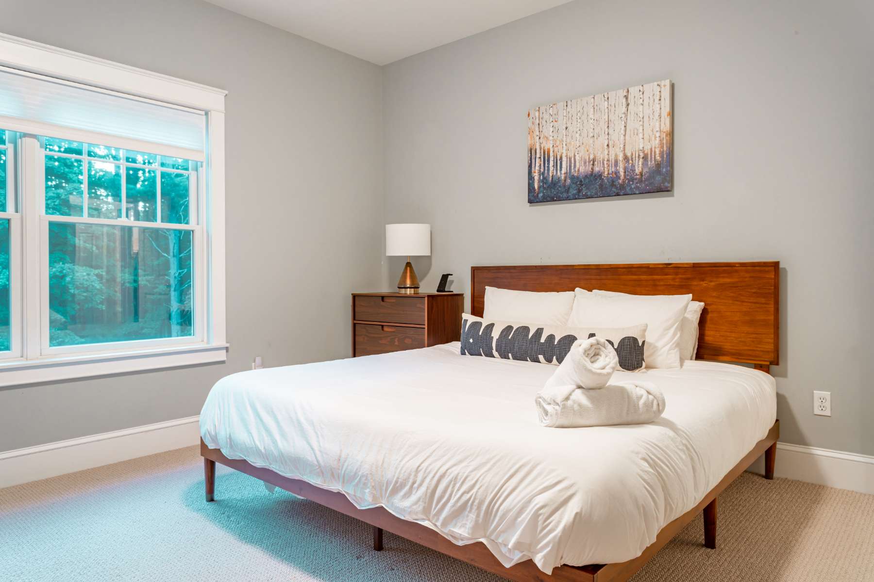 A cozy bedroom with a queen-sized bed featuring a wooden headboard and white bedding, accented with patterned pillows. A wooden nightstand with a lamp sits beside a large window offering natural light.