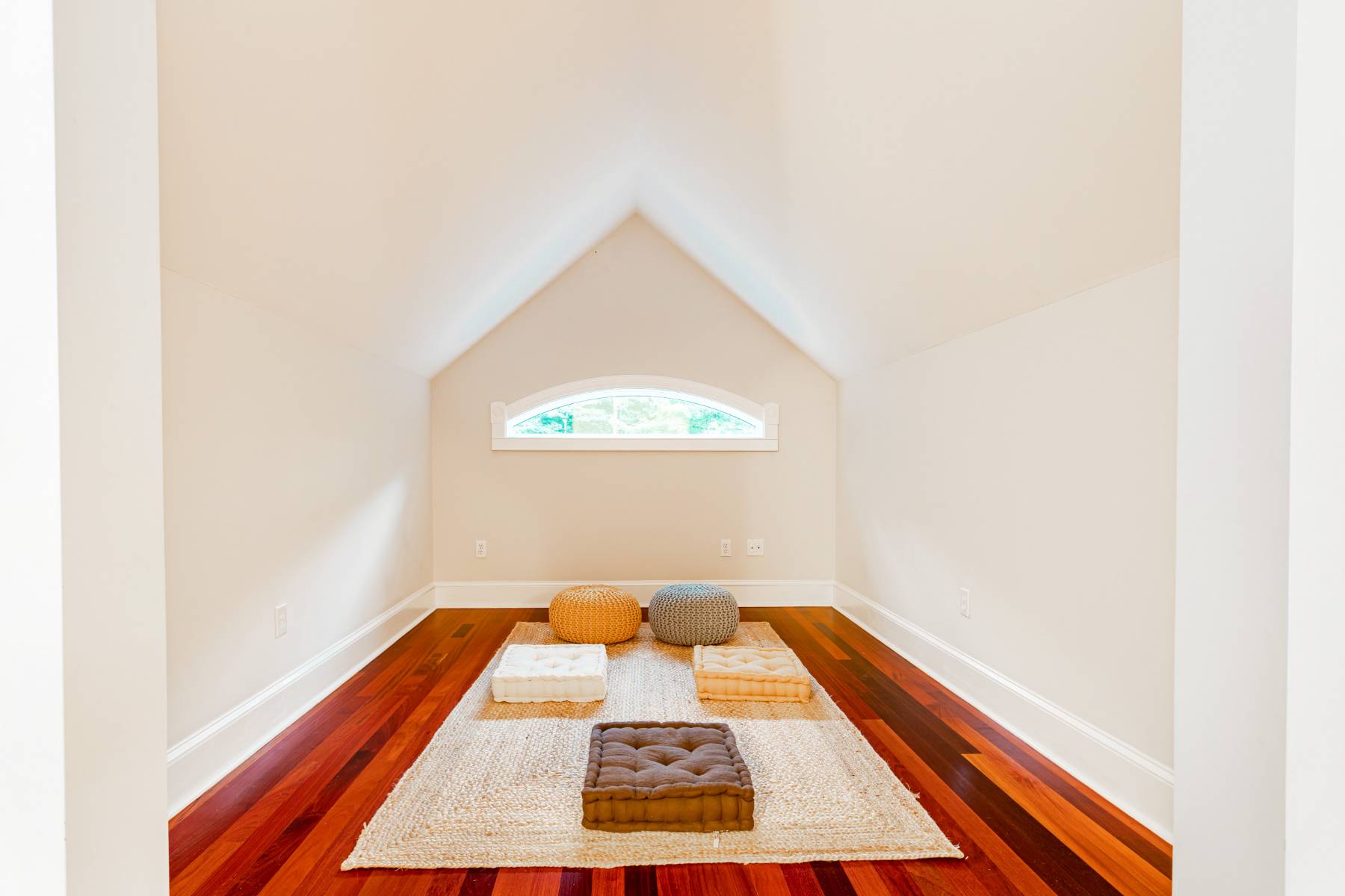 A serene attic space with a vaulted ceiling, a small arched window, and several floor cushions on a textured rug, creating a peaceful meditation or reading area on hardwood floors.