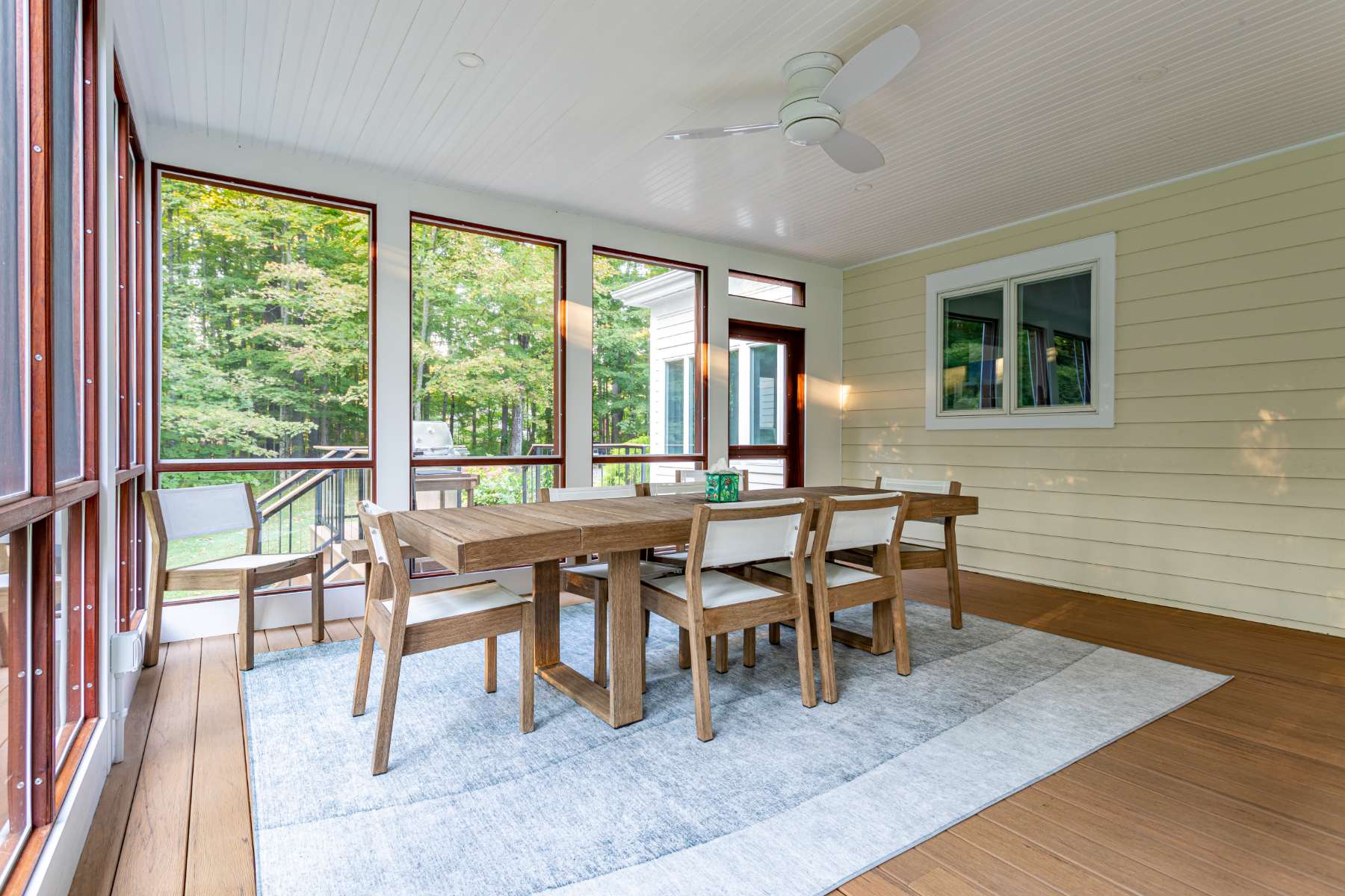 A screened-in dining area on a porch with light wood flooring and a white beadboard ceiling with a fan. A large wooden dining table with six chairs sits on a light blue and white rug, surrounded by views of the lush outdoors.