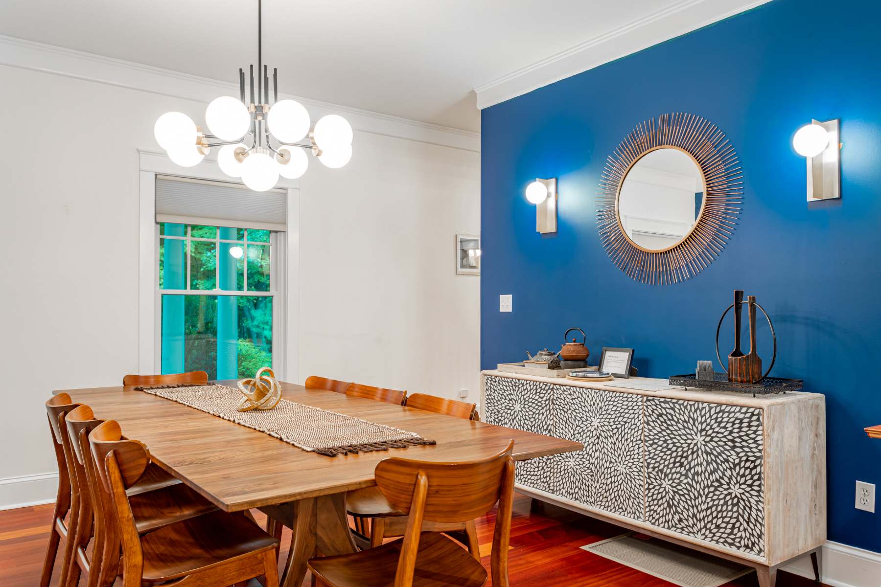 Elegant dining room with a large wooden table and chairs, set against a striking blue accent wall adorned with a sunburst mirror and sconces, complemented by a modern chandelier.