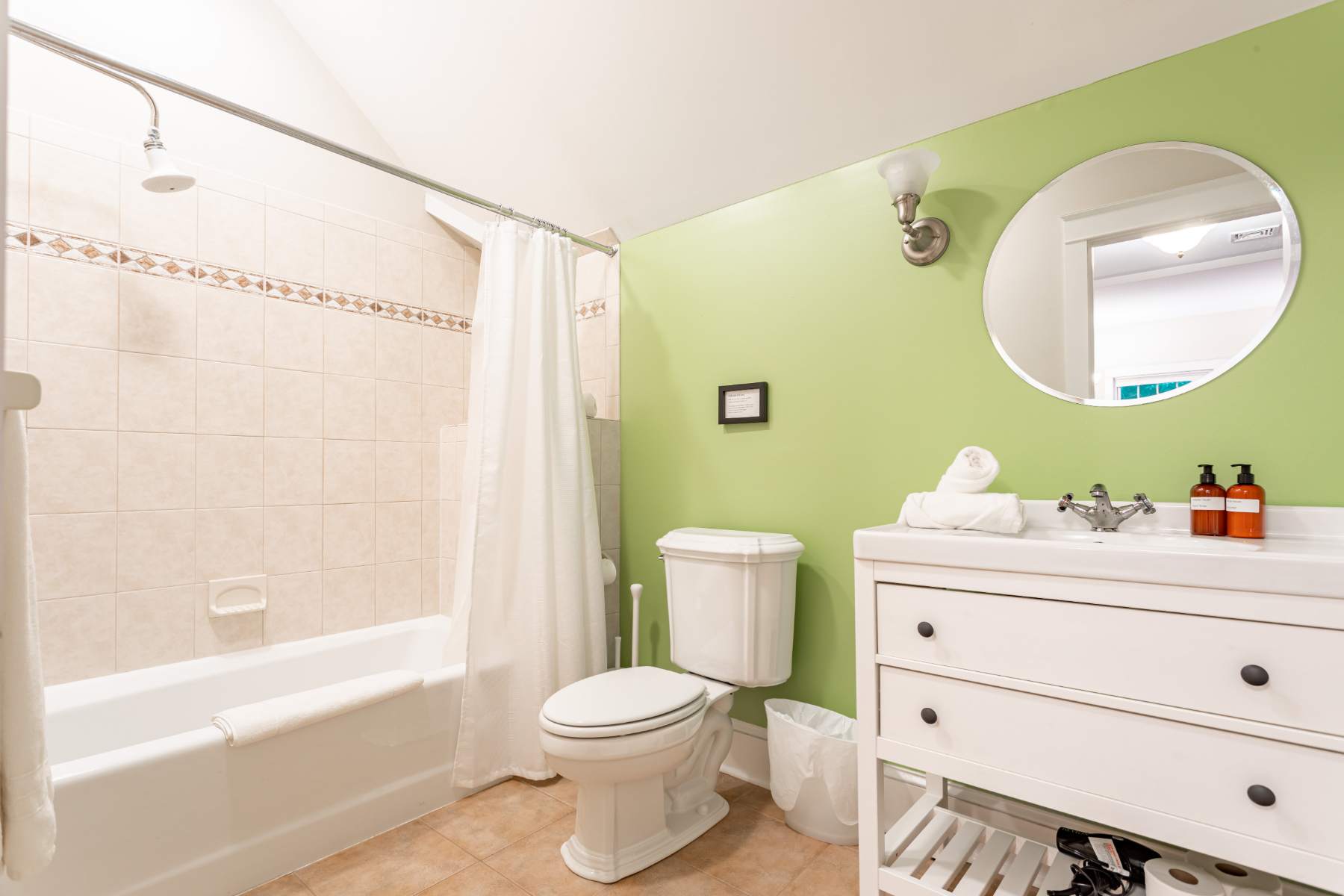 A bright bathroom featuring a white bathtub with a shower, a toilet, and a white vanity with a round mirror. The walls are painted a vibrant lime green, contrasting with the beige tiled shower and floor.