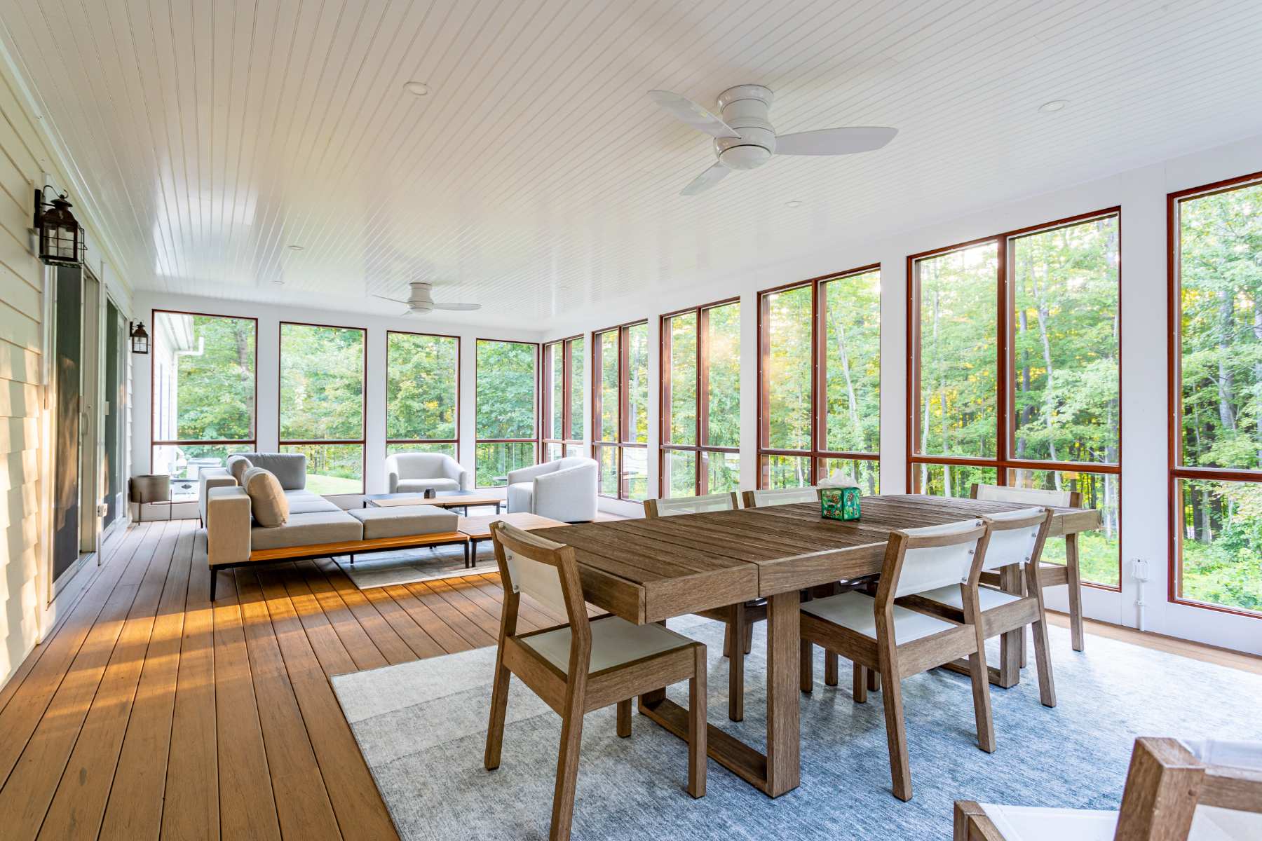 Expansive screened porch with light wood flooring and white paneled ceiling, furnished with a large gray sectional sofa, a wooden coffee table, and a dining area, all overlooking a lush forest.