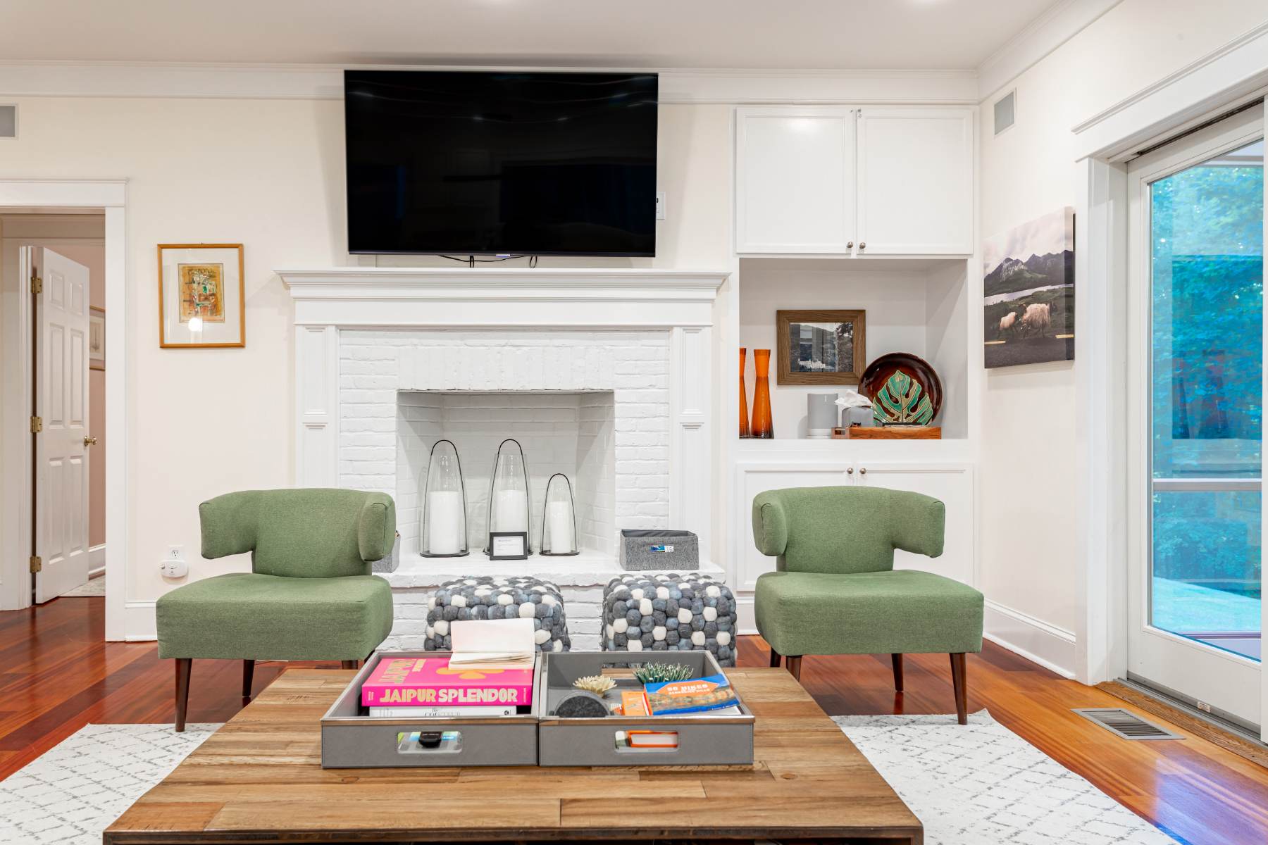 Cozy living room centered around a white brick fireplace with built-in shelving, featuring two green armchairs, patterned ottomans, and a large wall-mounted TV above the mantel.