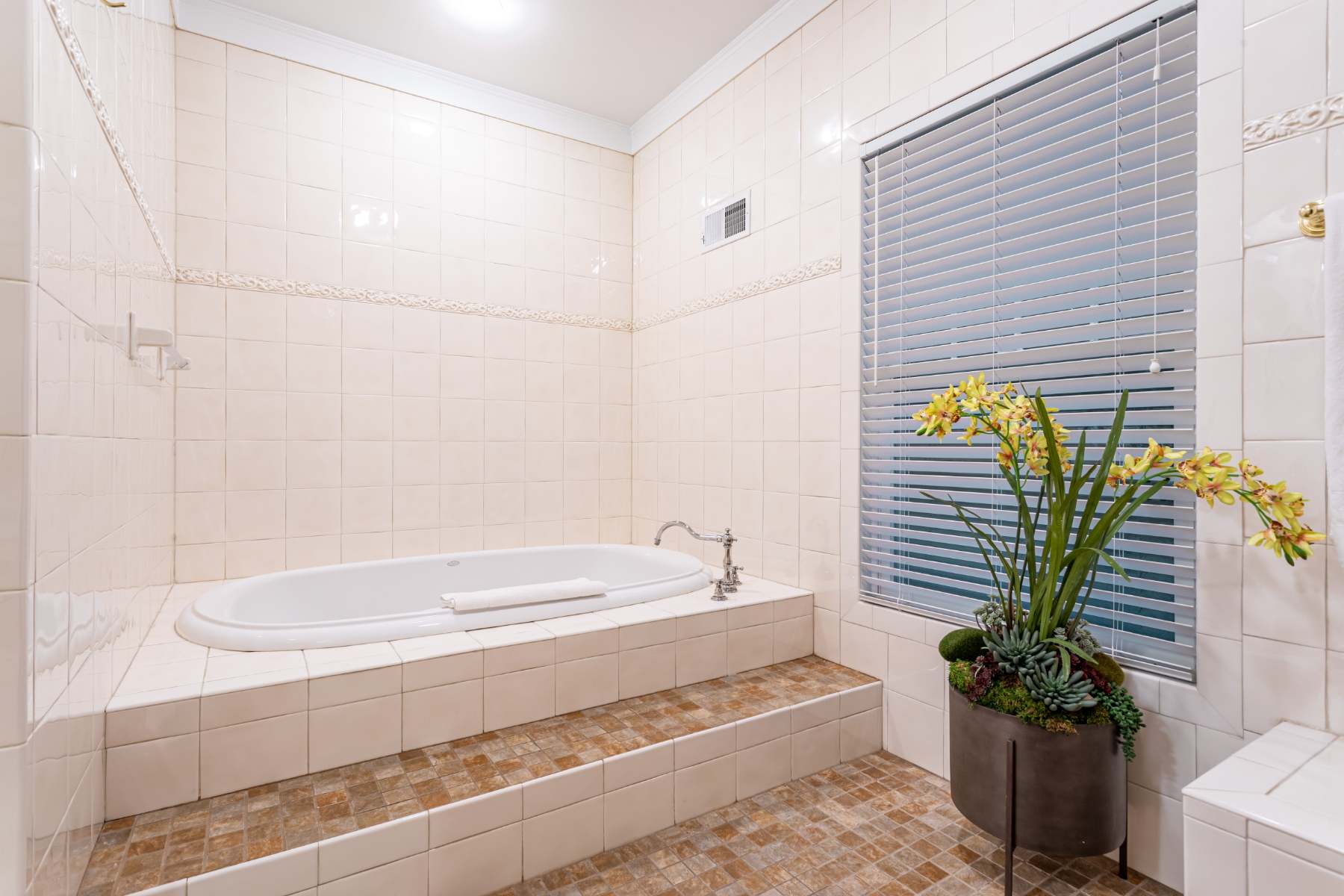 A spacious tiled bathroom featuring a built-in bathtub surrounded by light-colored tiles, with a large plant in a dark planter and a window with blinds, creating a serene bathing space.