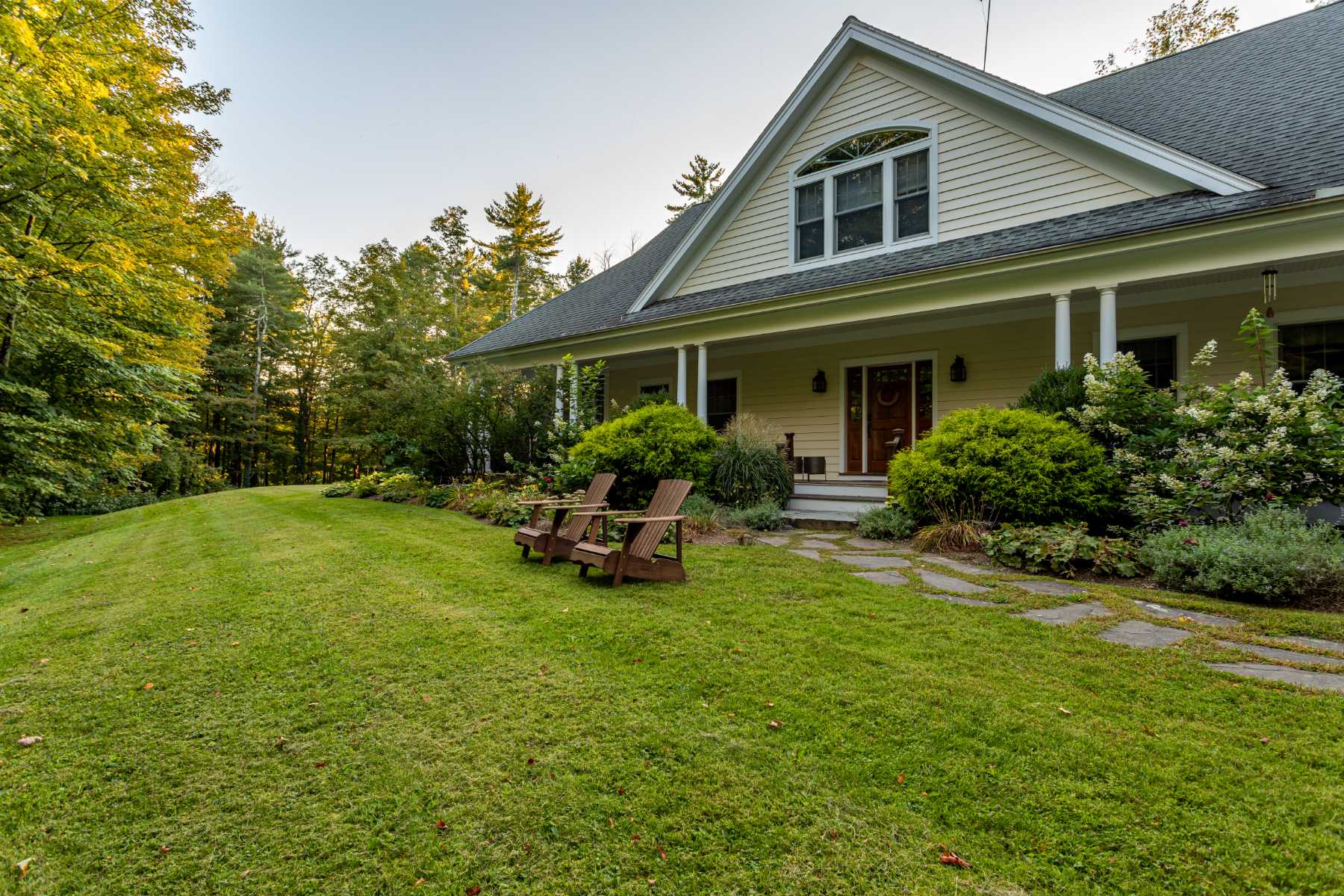 A ground-level view of the light yellow house's front facade, showcasing a wide green lawn and a stone path leading to the covered porch with white columns. Two wooden Adirondack chairs sit on the lawn, inviting relaxation.