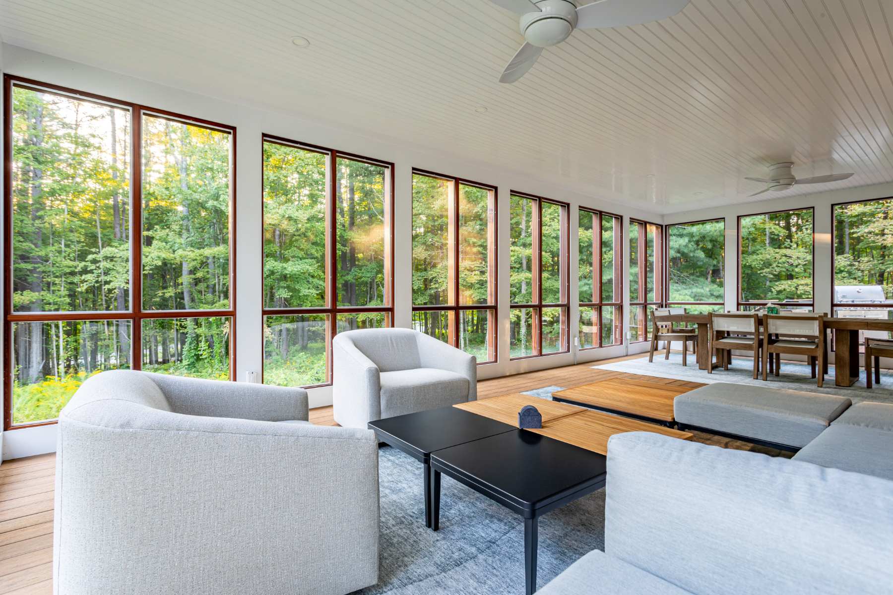 A bright, spacious screened porch with floor-to-ceiling windows offering extensive forest views, featuring a cozy seating area with light gray armchairs and a dining table in the background.