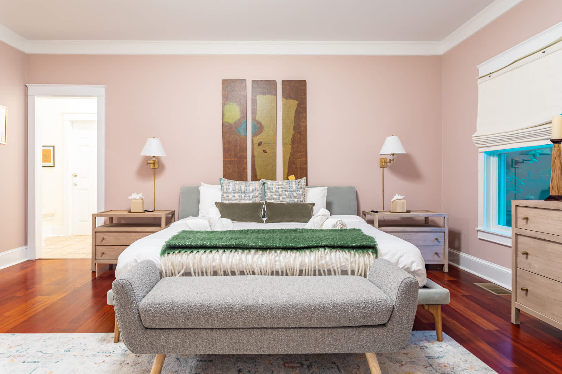 A spacious bedroom with light pink walls, a large bed with a grey headboard, two wooden nightstands with lamps, and a grey bench at the foot of the bed, all on polished hardwood floors.