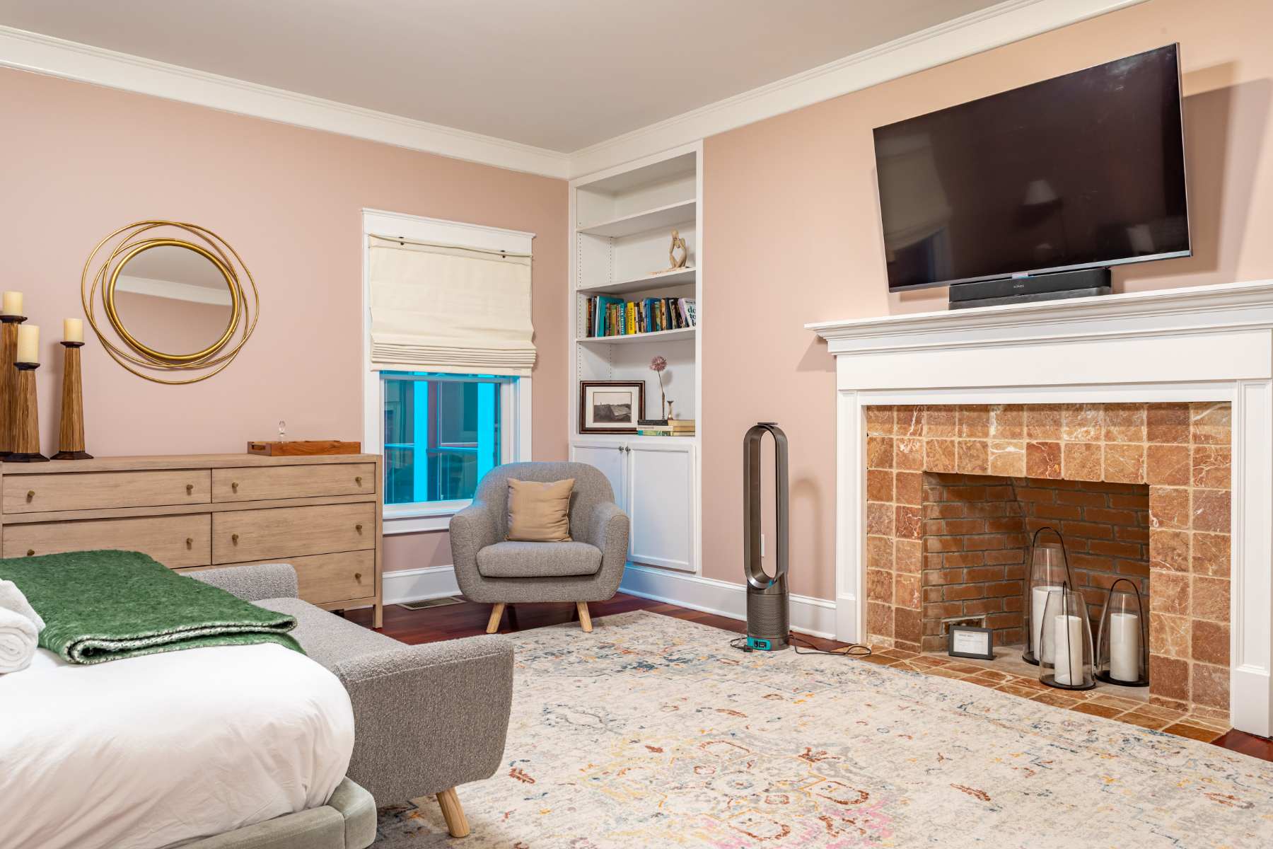 A cozy corner of a bedroom featuring a light pink wall, a large wall-mounted TV above a fireplace with brick surround, a built-in bookshelf, and a grey armchair on an area rug.