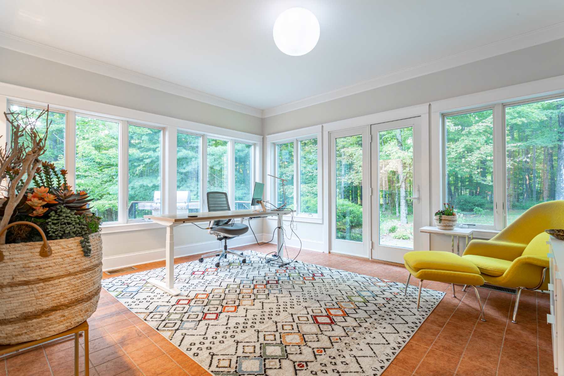 Bright home office with multiple large windows showcasing a lush green forest, featuring a modern desk with two chairs, a vibrant patterned area rug, and a yellow accent armchair.