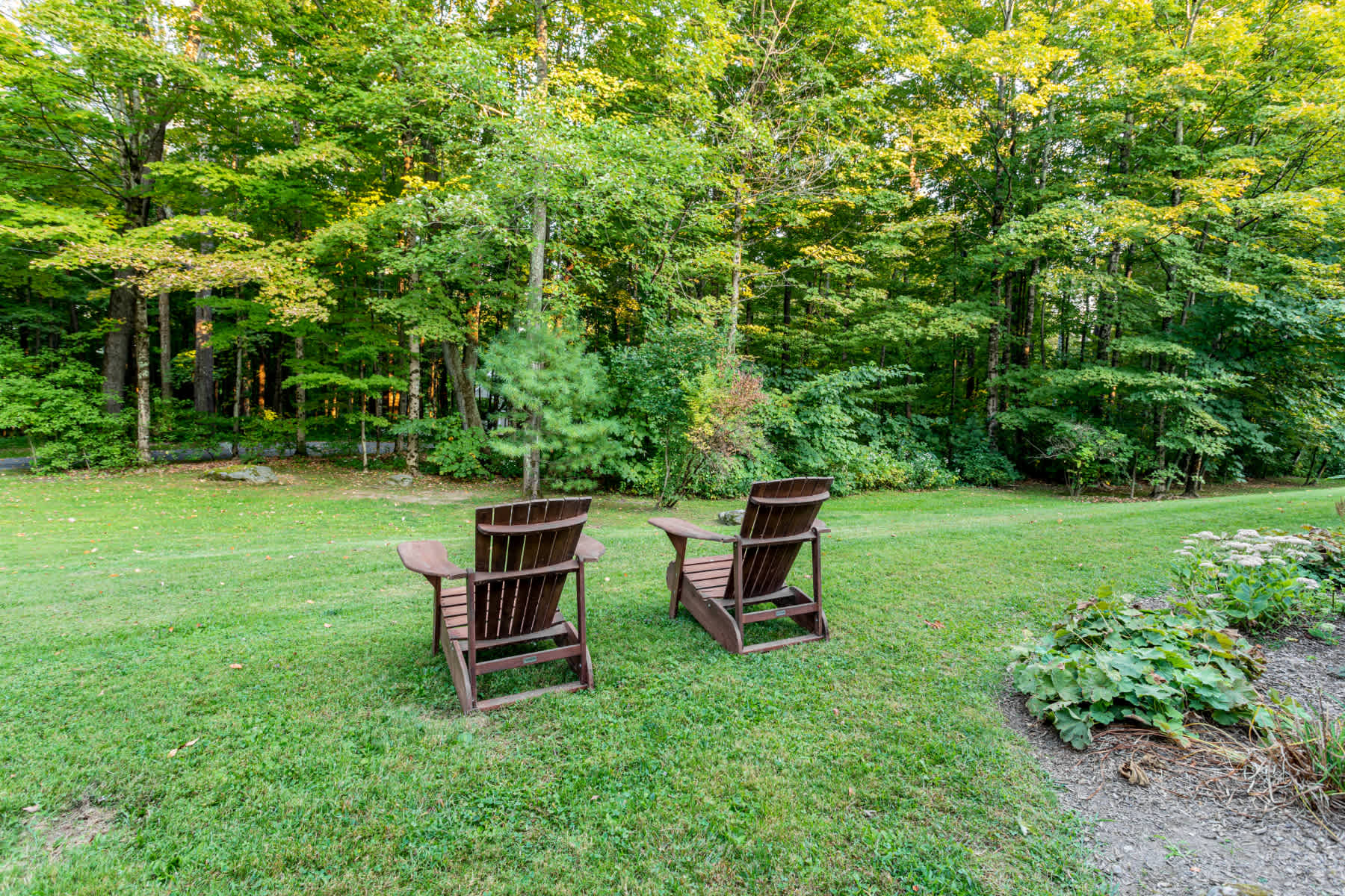 A serene outdoor scene featuring two wooden Adirondack chairs placed on a well-maintained green lawn, facing a dense backdrop of mature trees. The setting suggests a peaceful garden or backyard area.