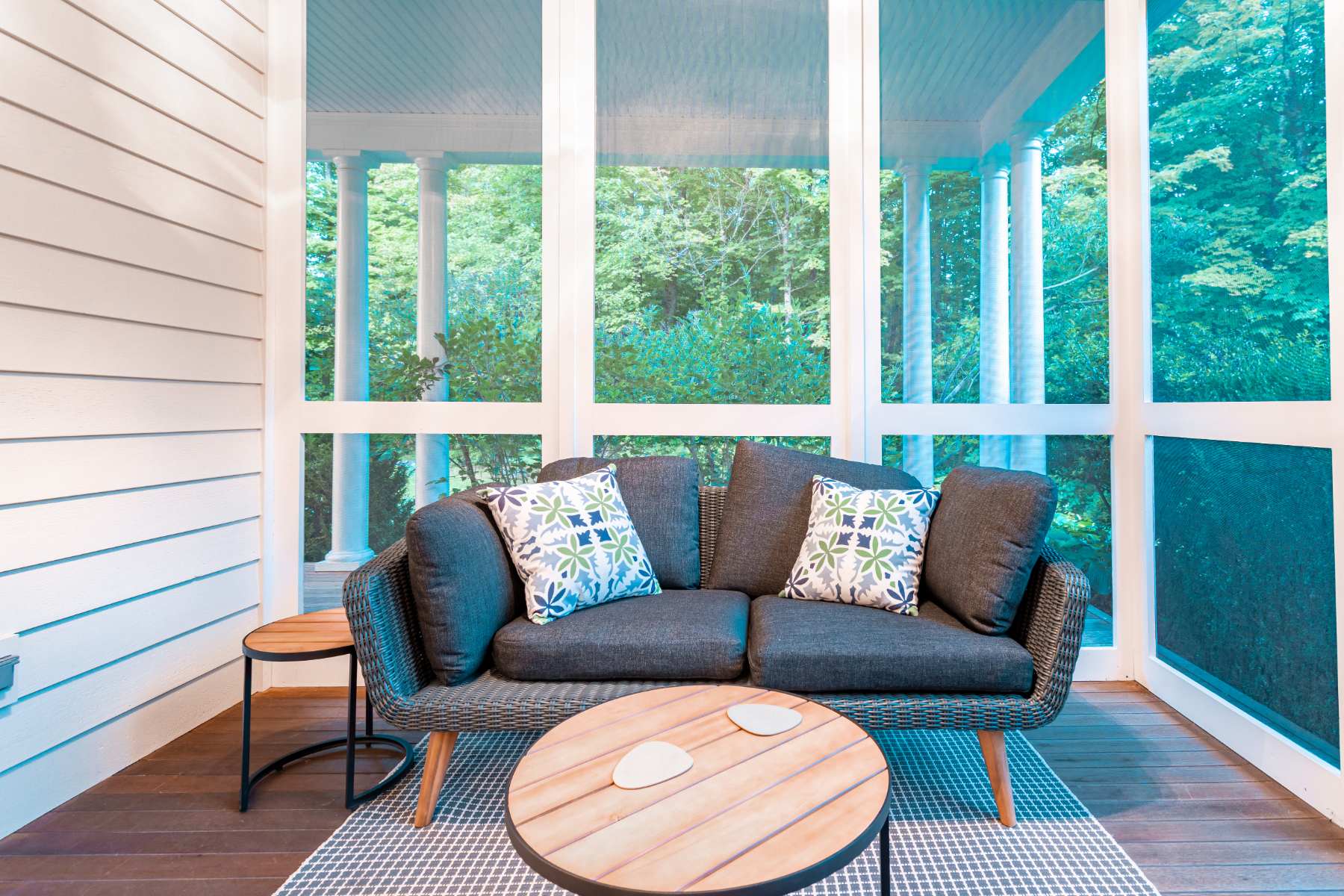 A screened porch with a dark grey outdoor sofa, two round wooden side tables, and a round wooden coffee table on a patterned rug, offering views of lush green trees.