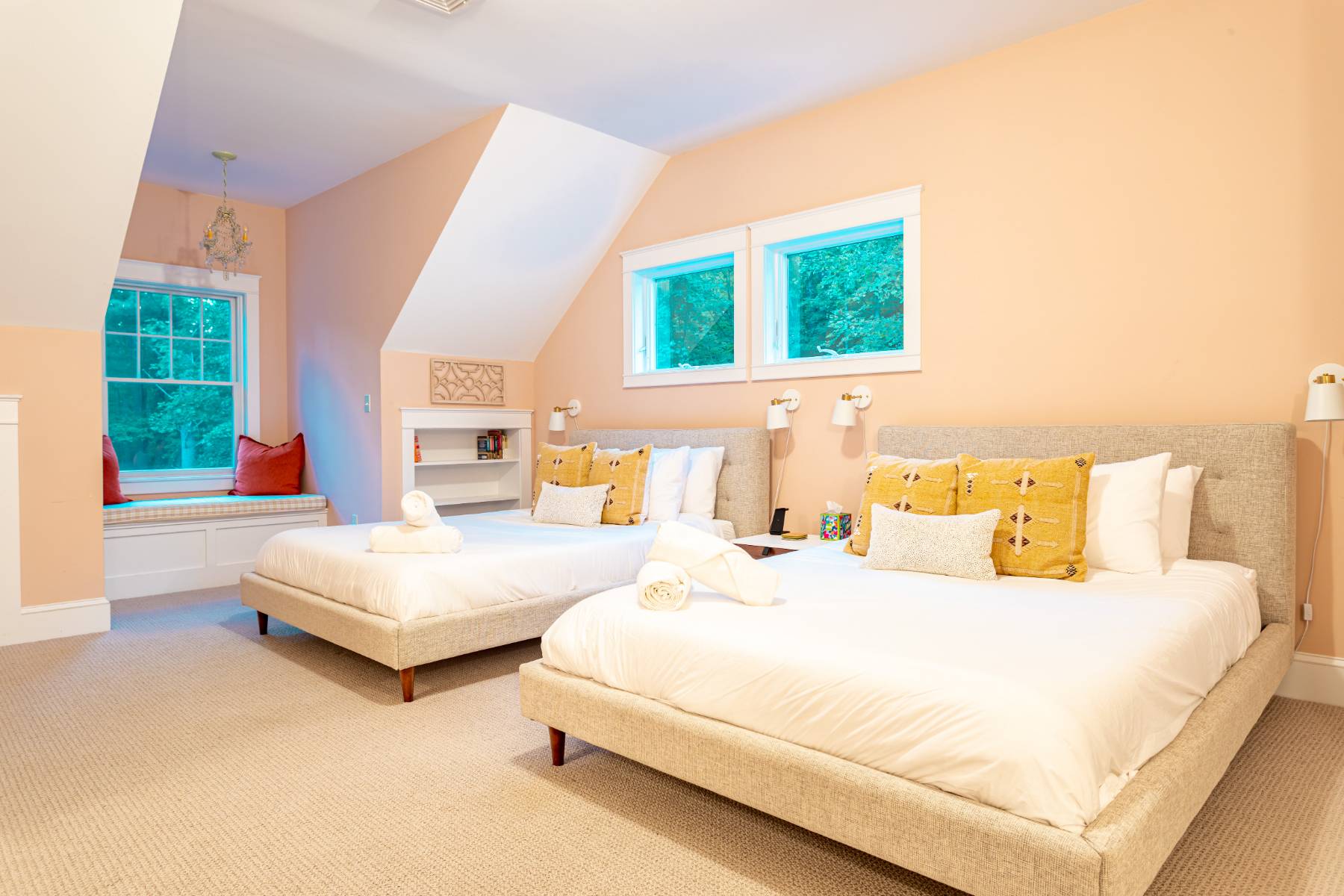 A spacious bedroom with two queen-sized beds, each with light grey upholstered frames and yellow accent pillows. The room features sloped ceilings, multiple windows, and a built-in window seat with red cushions.