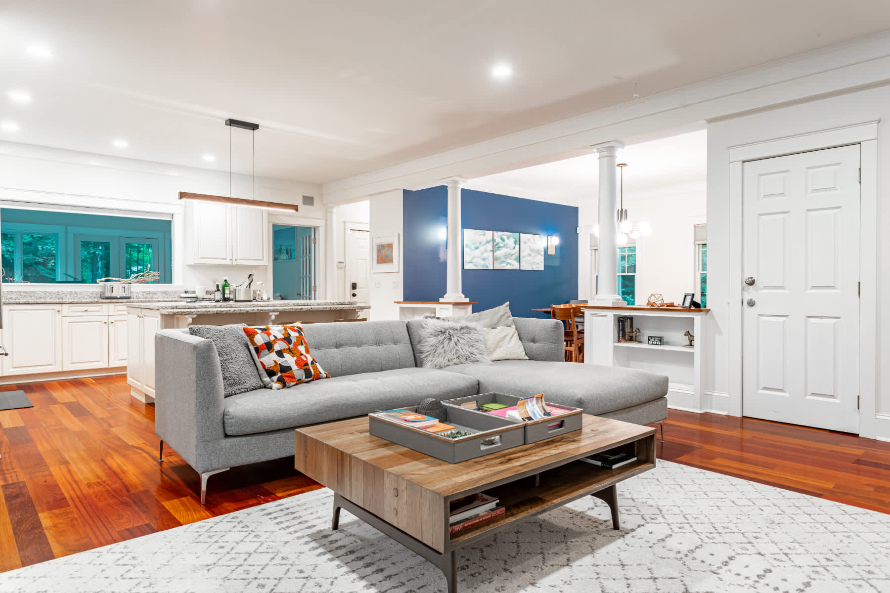 Open-concept living space featuring a gray sectional sofa and a wooden coffee table on a light rug, with a view into the modern white kitchen and a dining area with a bold blue accent wall.