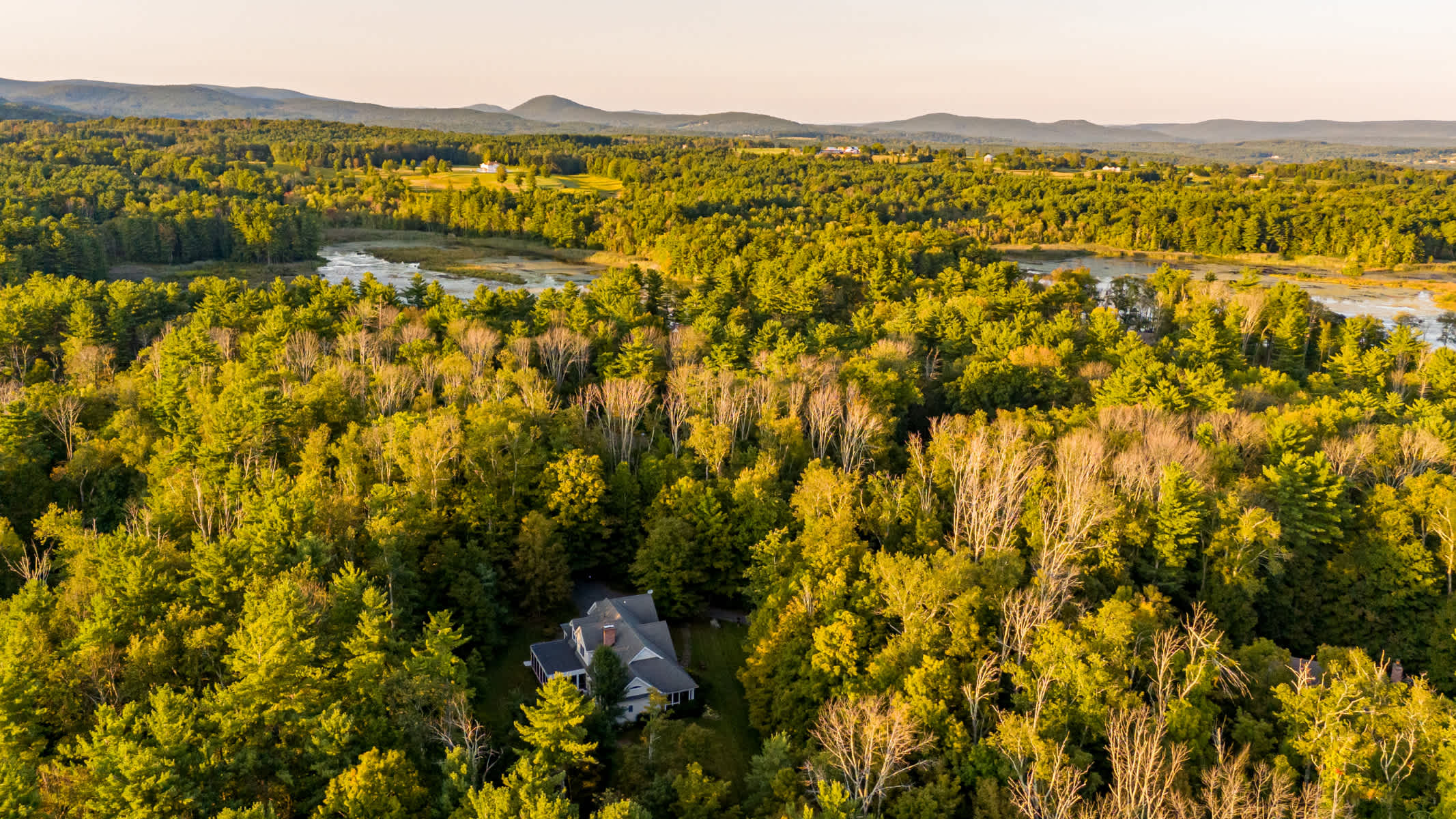An aerial view showcases a large house nestled deep within a dense, vibrant forest displaying early autumn colors, with a winding river or marsh visible in the mid-ground, and rolling mountains under a clear sky in the distance.