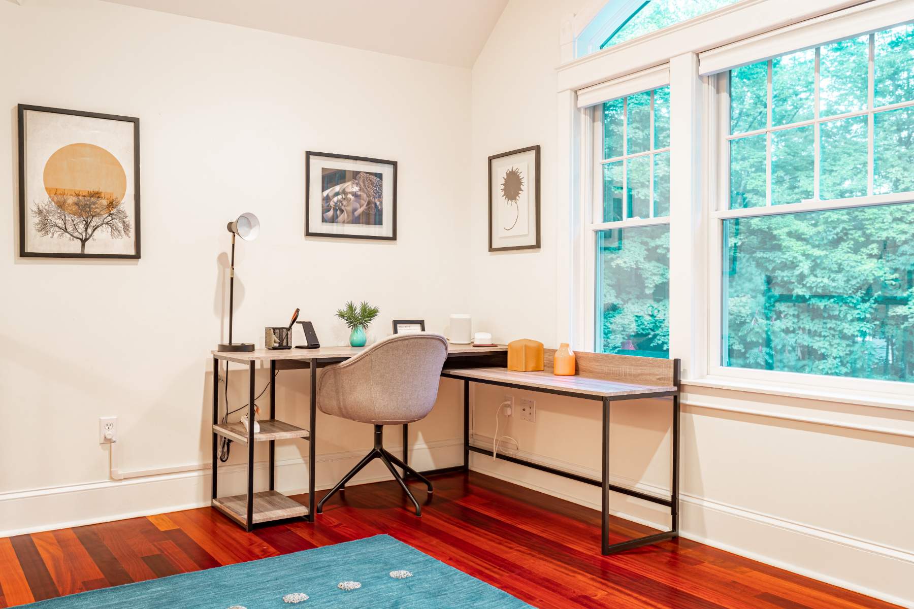 A bright office space with a modern L-shaped desk, a grey office chair, and large windows offering views of lush greenery, all set on polished hardwood floors with contemporary artwork.