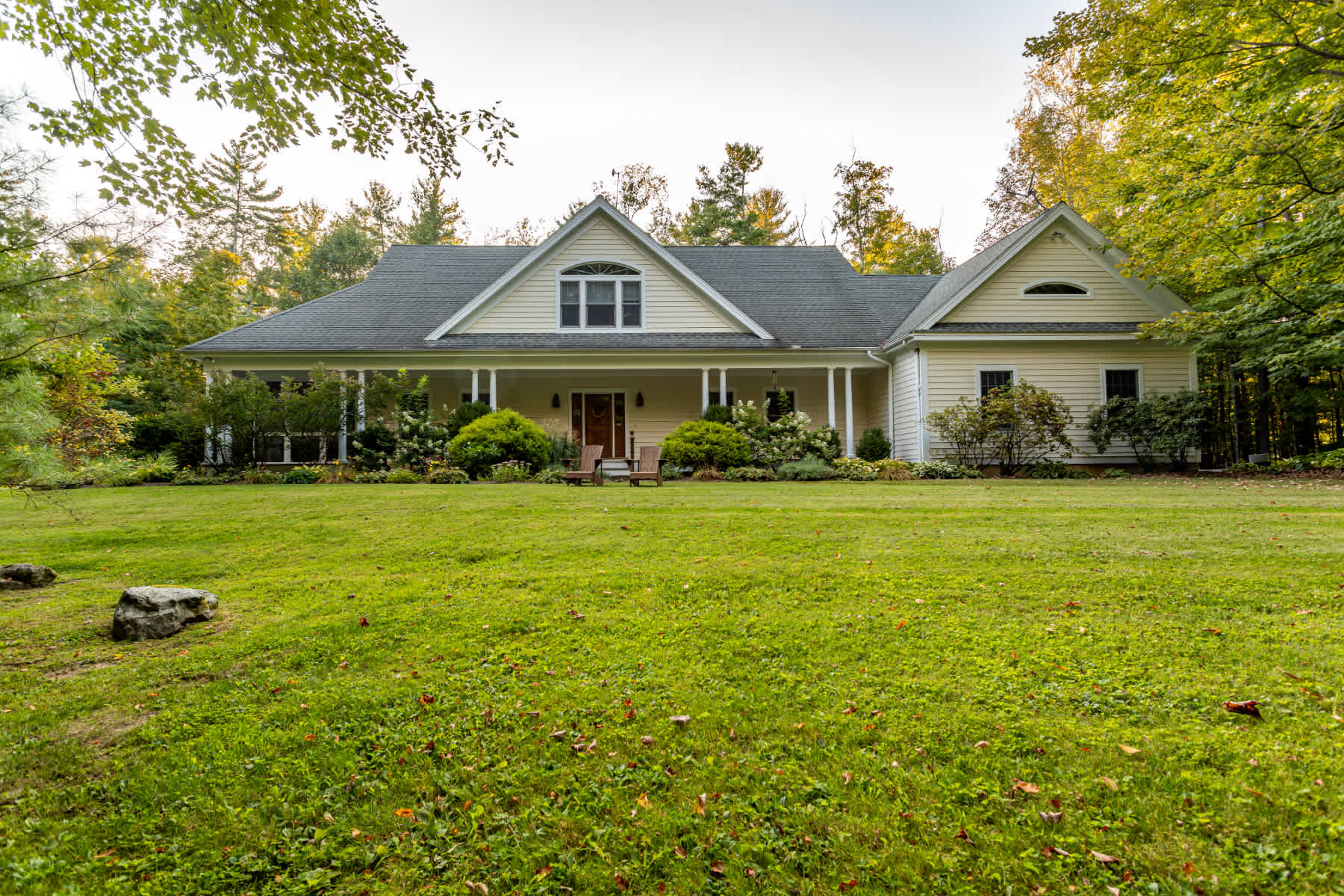 The front exterior of a charming light-colored house with a wide covered porch supported by white columns. The house is set on a large green lawn, surrounded by mature trees and landscaping.