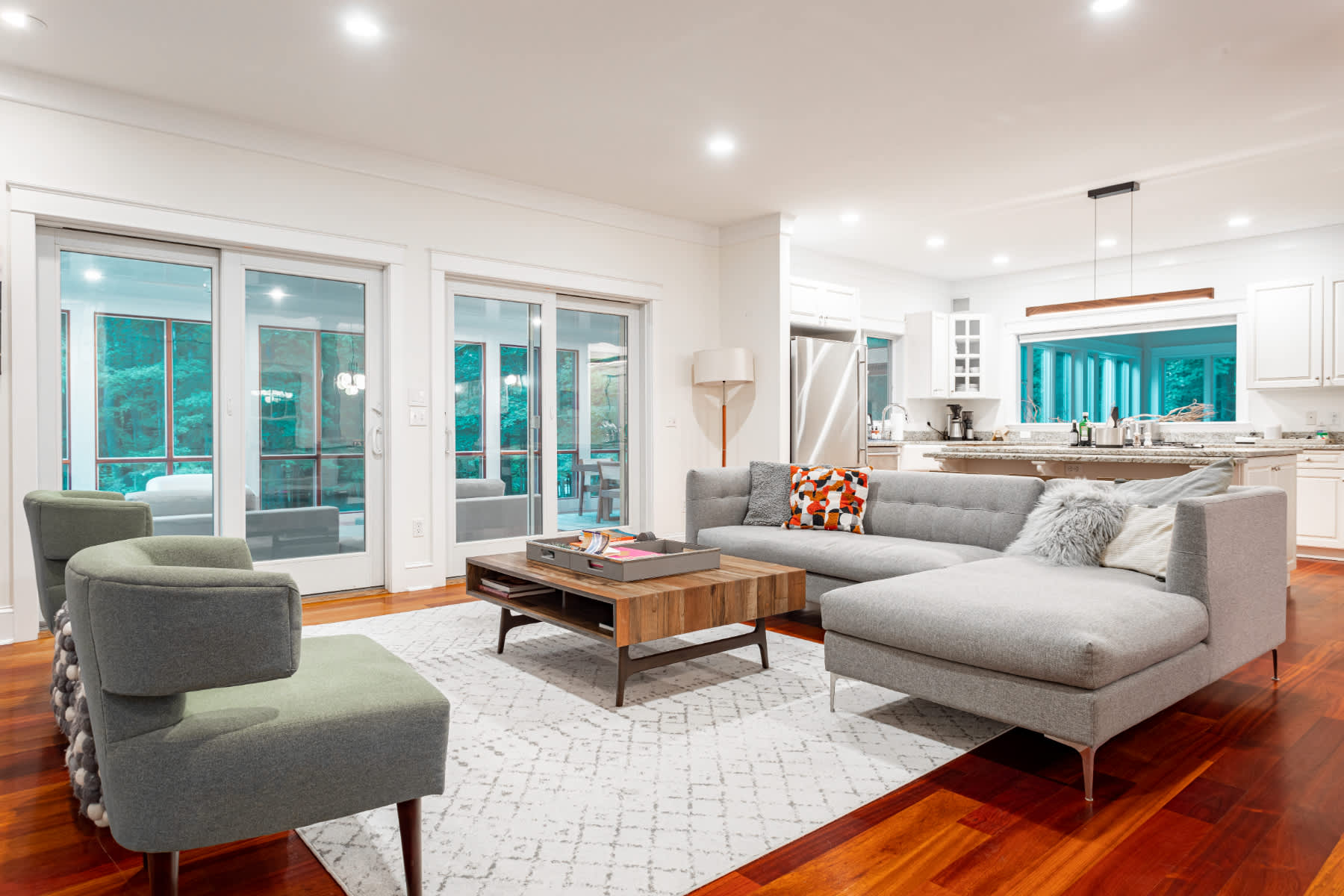 A bright living room with rich reddish-brown hardwood floors and white walls, featuring a large gray sectional sofa and two green armchairs. Sliding glass doors open to a screened porch, and a modern kitchen is visible in the background.