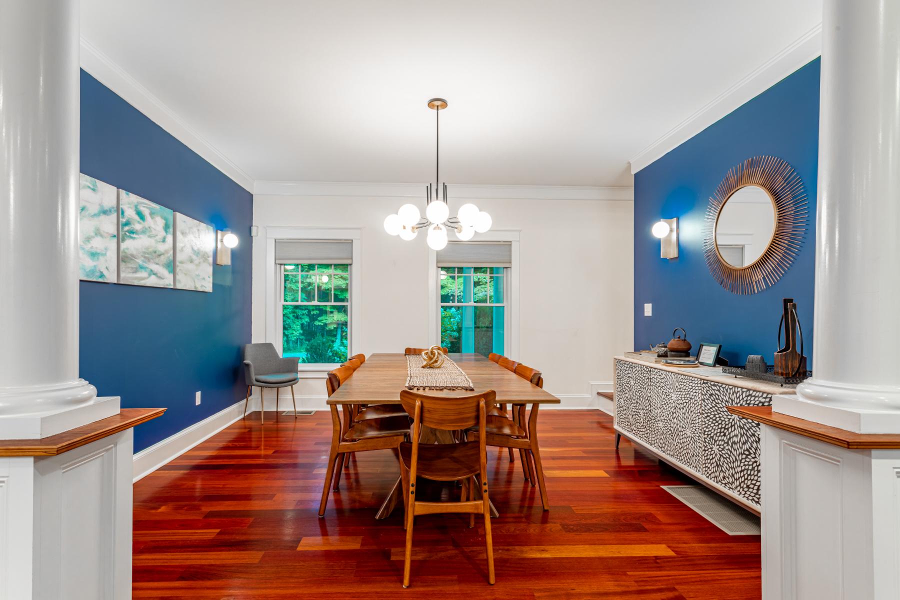 A formal dining room with rich reddish-brown hardwood floors and a striking accent wall in deep blue. A long wooden dining table is surrounded by six wooden chairs, illuminated by a modern chandelier.