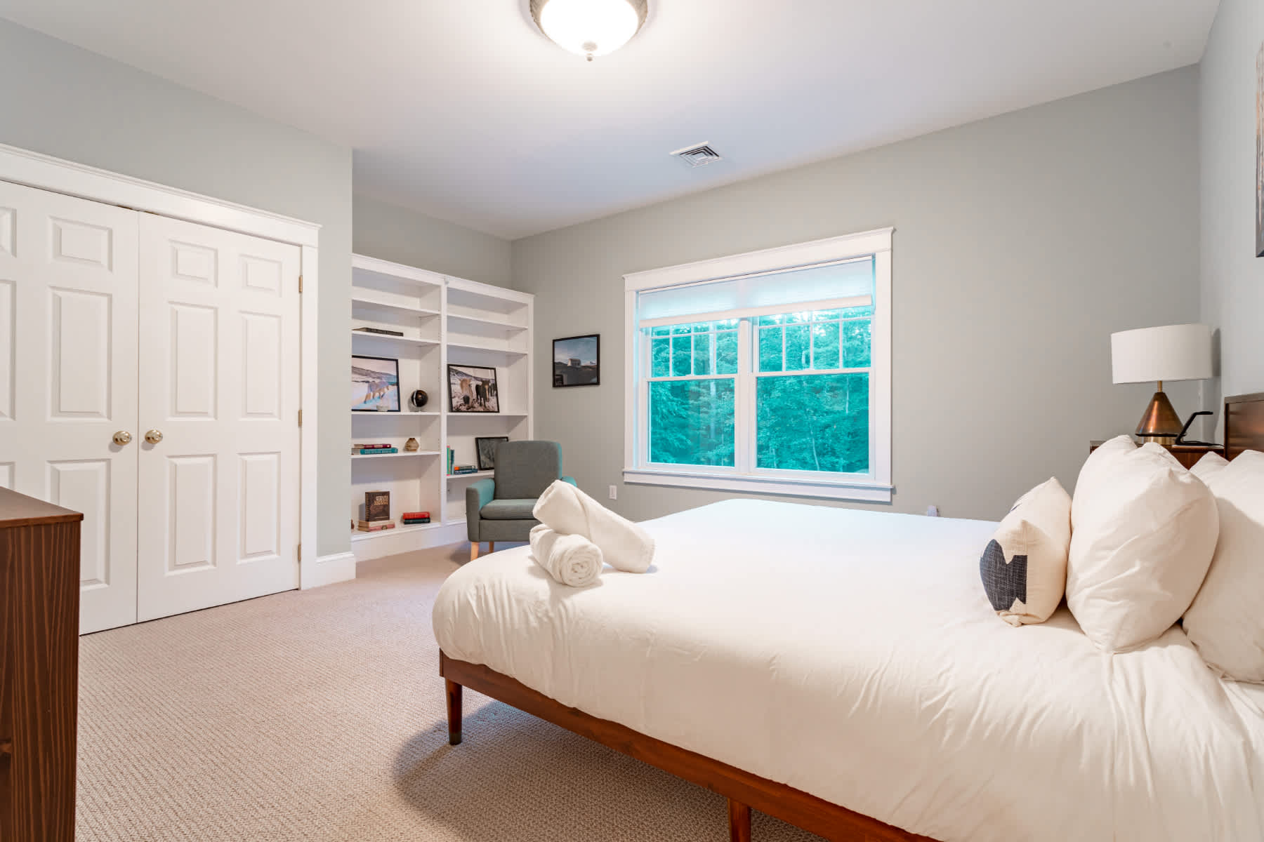 View of a bedroom with a queen bed, white bedding, and a wooden headboard. The room includes a white built-in bookshelf, a grey armchair, and a double-door closet, with a large window overlooking greenery.