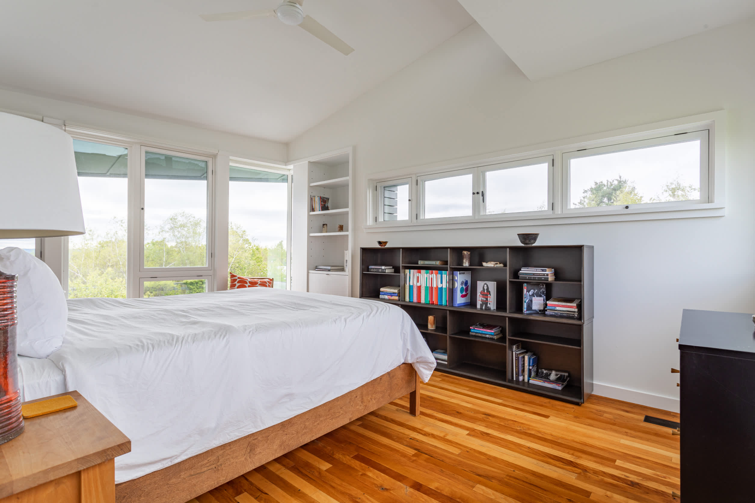 Bright bedroom with a white bed, light wood frame, and large windows offering views of greenery. A dark wood bookshelf is built into the wall, and the room features warm hardwood floors and a vaulted ceiling.