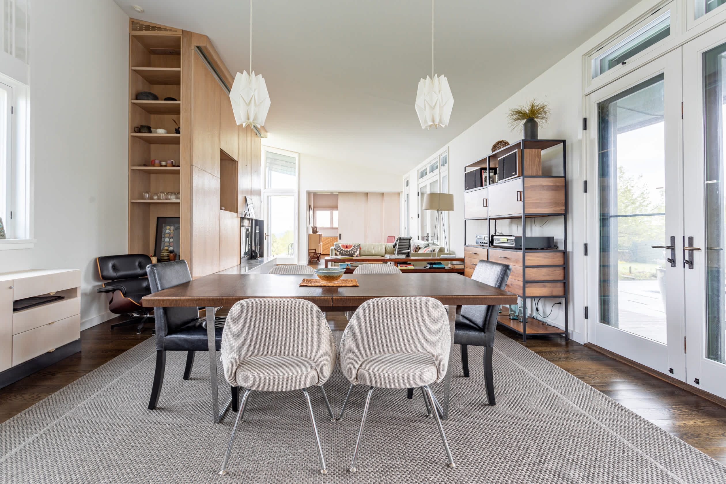 Contemporary dining area with a dark wood table and mixed chairs on a light grey rug, flanked by a tall wooden shelving unit and large glass doors.