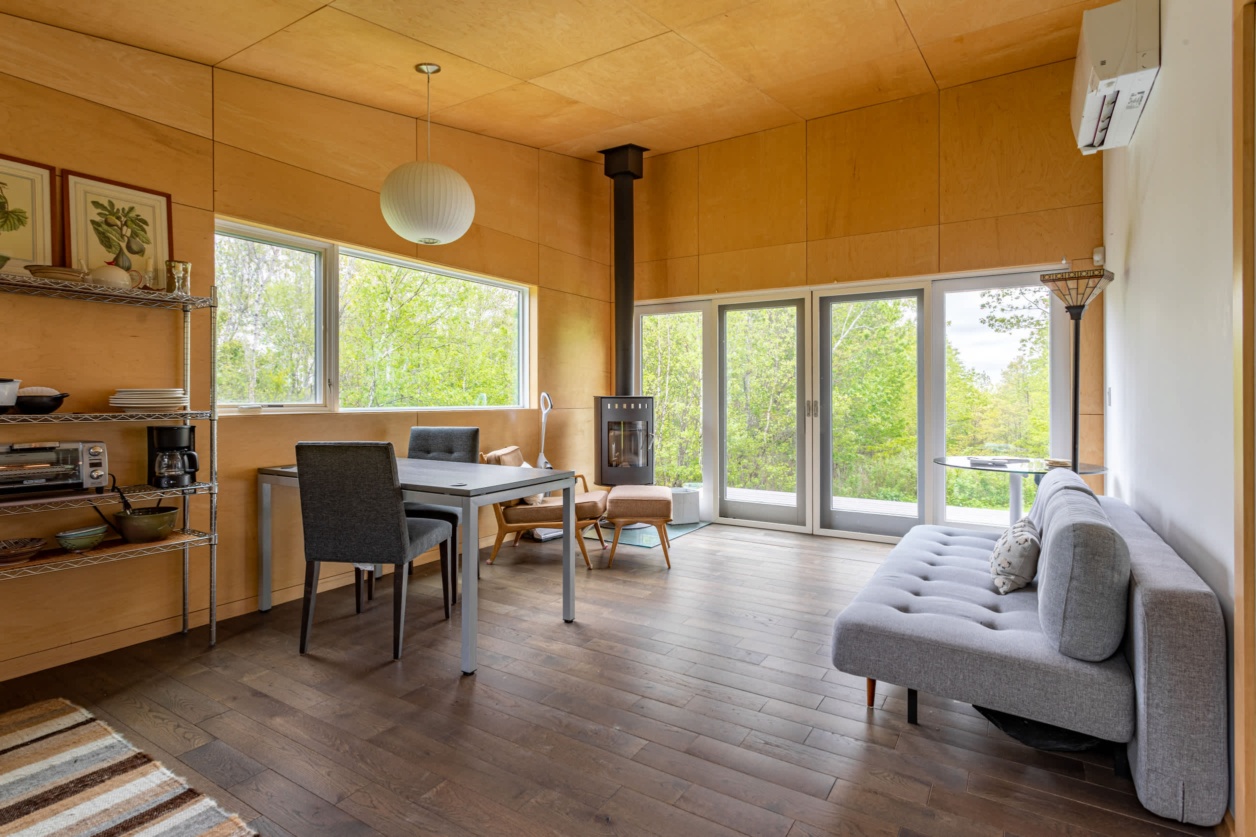 Open-concept living and dining area with light wood paneling, featuring a gray sofa, a dining table with chairs, and a wood-burning stove. Large windows offer views of the surrounding trees.