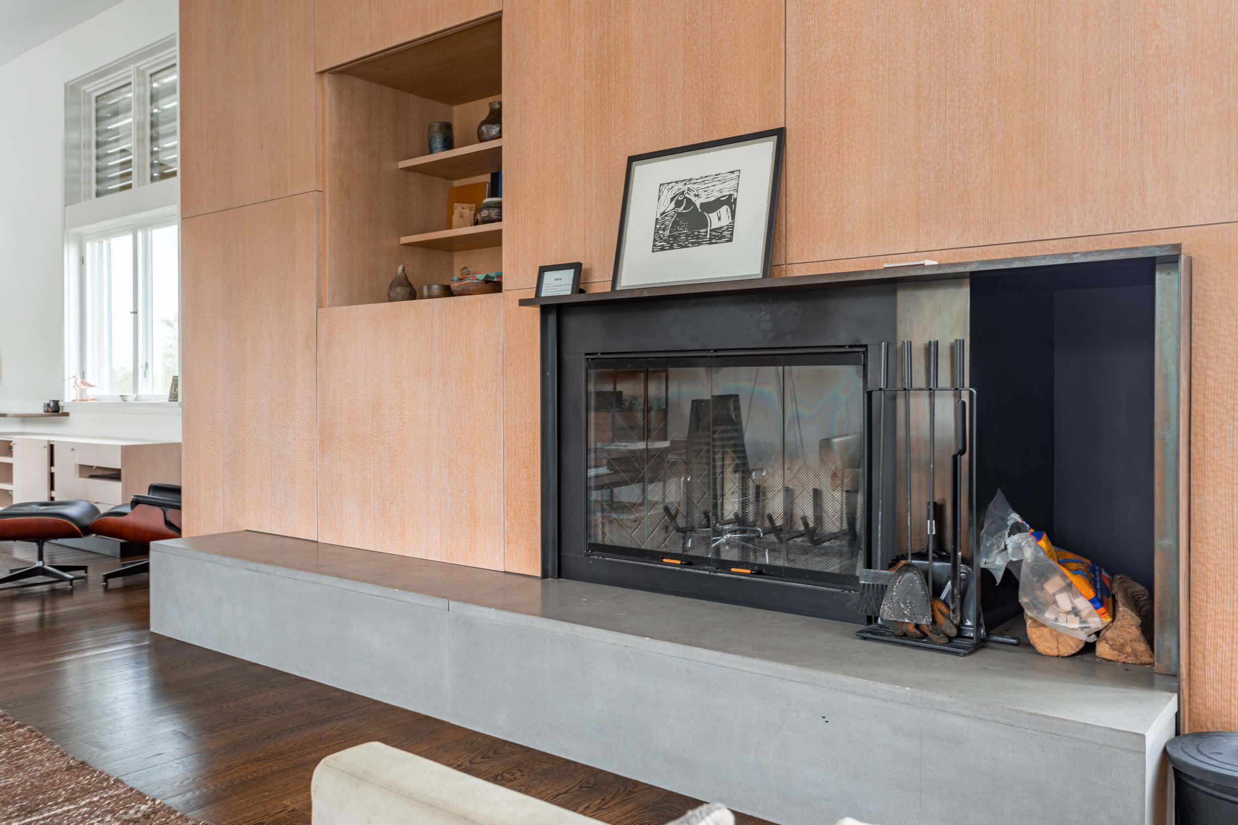 Modern fireplace with a black insert and concrete hearth, integrated into a light wood paneled wall with built-in shelving. Fireplace tools and wood are visible.