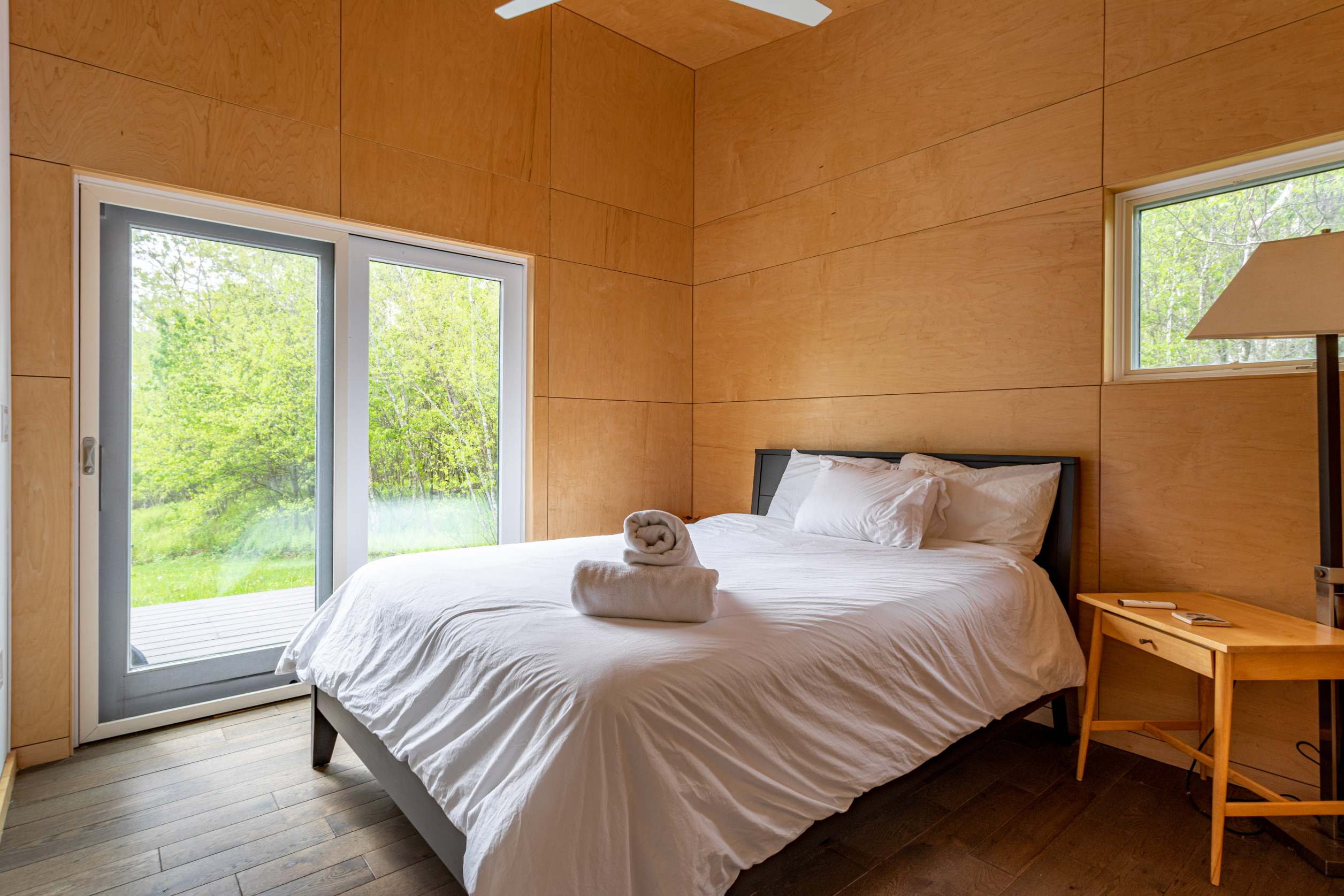 A bright bedroom with light wood paneling, a bed with white bedding and towels, and large glass doors leading to an outdoor area with lush greenery. A wooden nightstand and floor lamp are visible on the right.