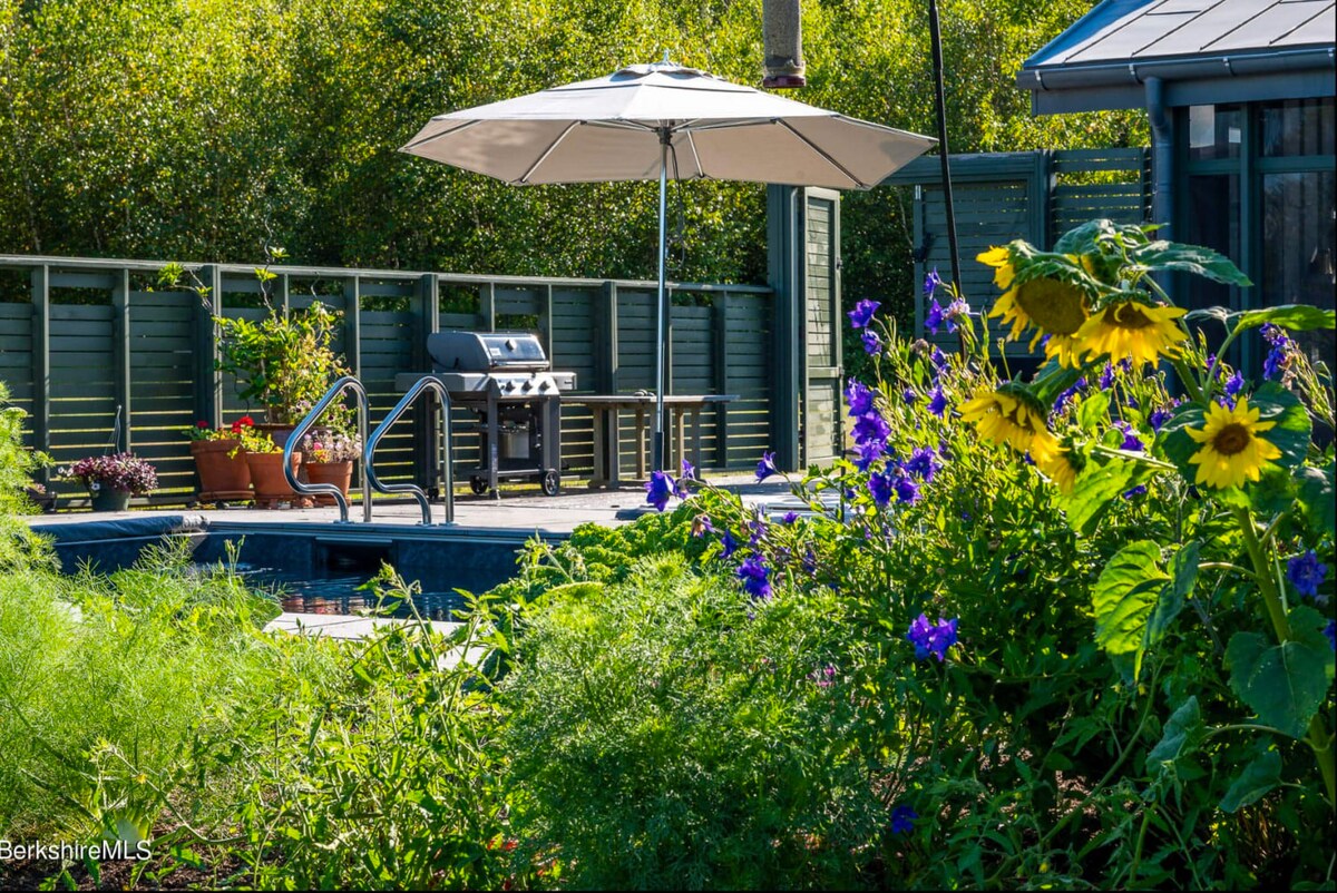 A vibrant garden area with sunflowers and purple flowers, set against a backdrop of lush green foliage. A BBQ grill and a white patio umbrella are visible in the background, near a swimming pool.