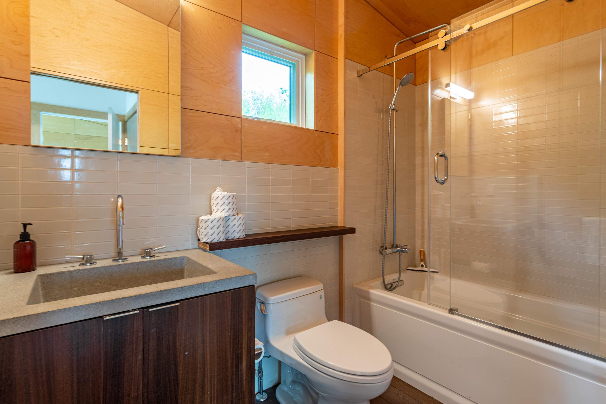 A modern bathroom with light-colored subway tiles, a concrete sink vanity with dark wood cabinets, and a toilet. A glass-enclosed shower/tub combo is visible on the right, with a small window above the toilet.