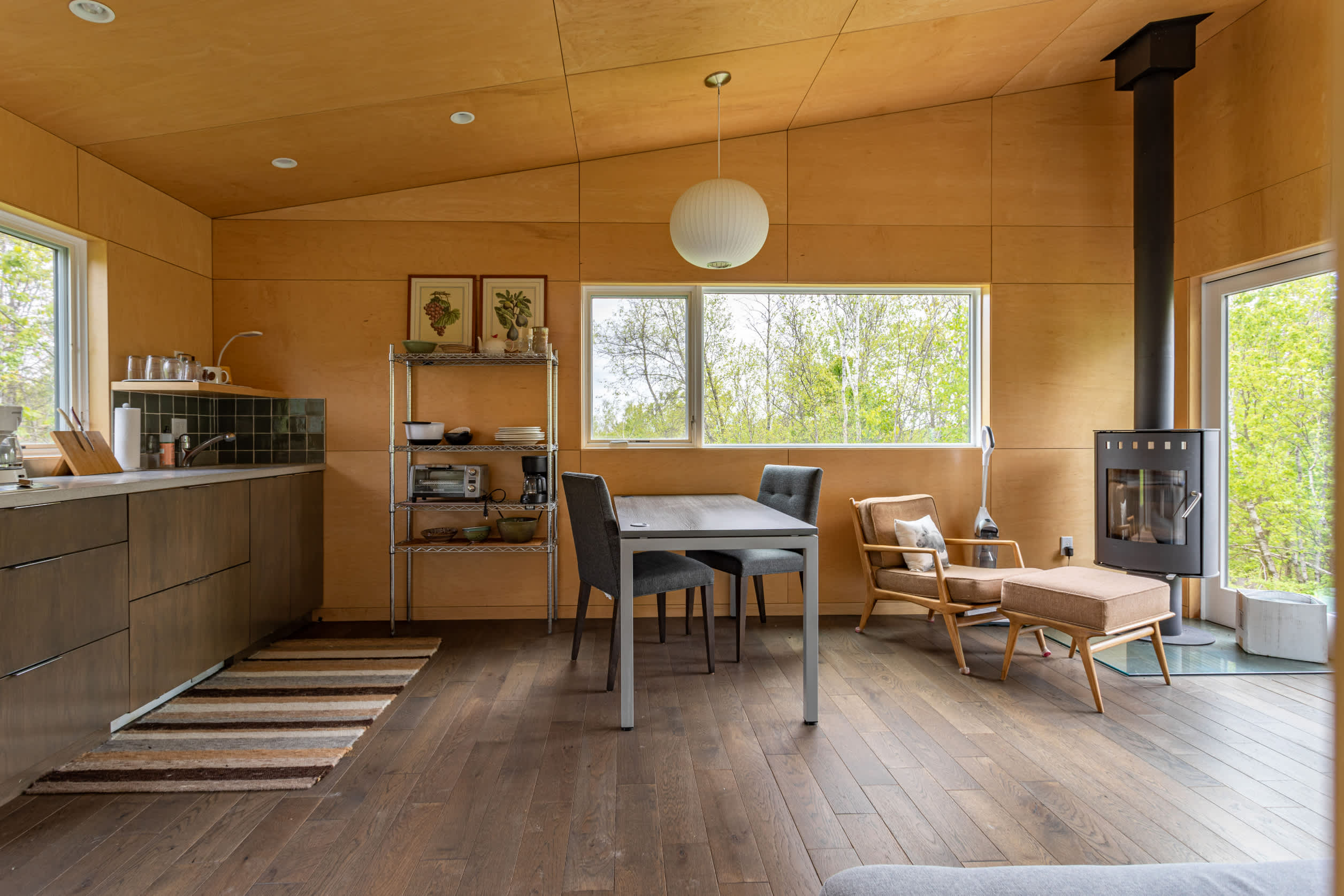 Cozy interior space with light wood paneling on walls and ceiling, featuring a compact kitchen area with dark cabinets and a dining table with chairs. A wood-burning stove and a lounge chair are also visible.