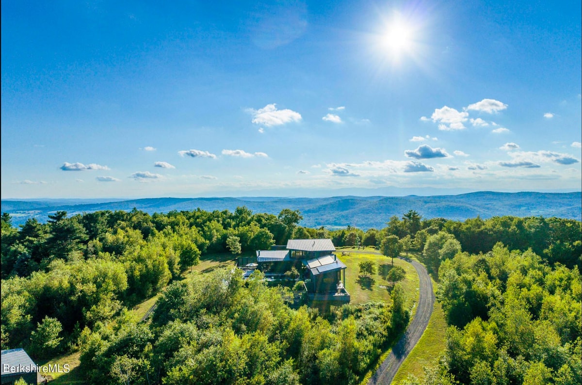 An aerial view of the modern green house nestled among abundant green trees, showcasing its unique architecture and surrounding natural landscape. A winding driveway leads to the property under a bright sunny sky.