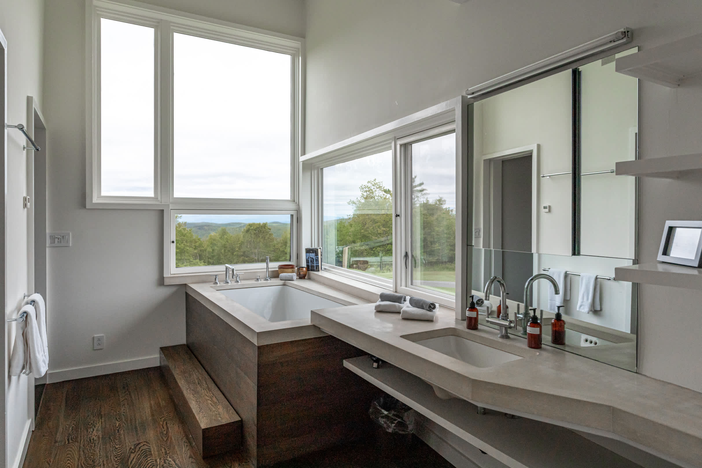Bright, modern bathroom with a built-in bathtub surrounded by wood paneling and a large window offering scenic views. A double vanity with concrete countertops and sleek faucets is also present.