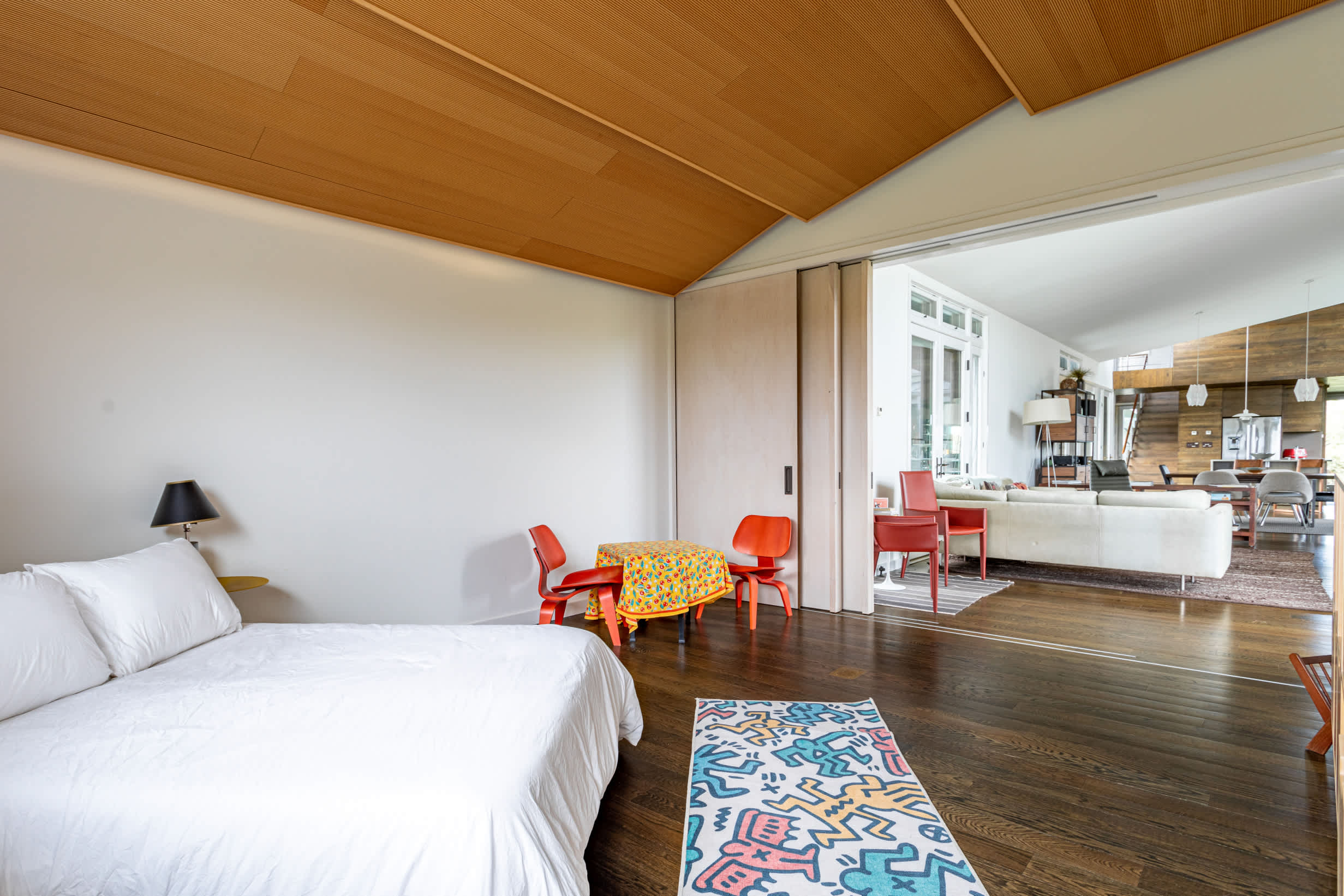 Wide shot of a contemporary bedroom with dark wood floors and a colorful rug, showing the open connection to a bright living area through large sliding doors. A small red table and chairs are positioned near the entrance.