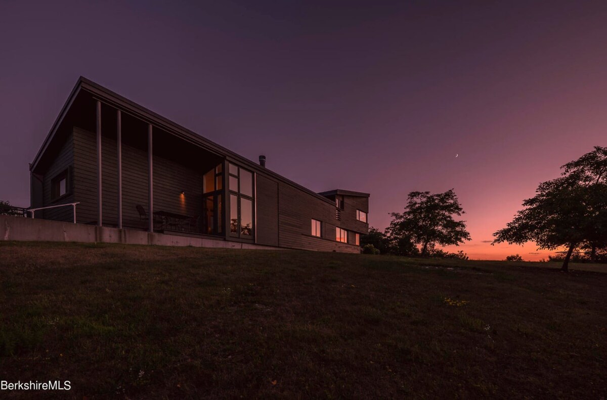 Modern dark-sided house with large windows and a sloped roof, illuminated by interior lights, set against a dramatic purple and orange twilight sky with a crescent moon. The house is situated on a gentle grassy slope, surrounded by trees.