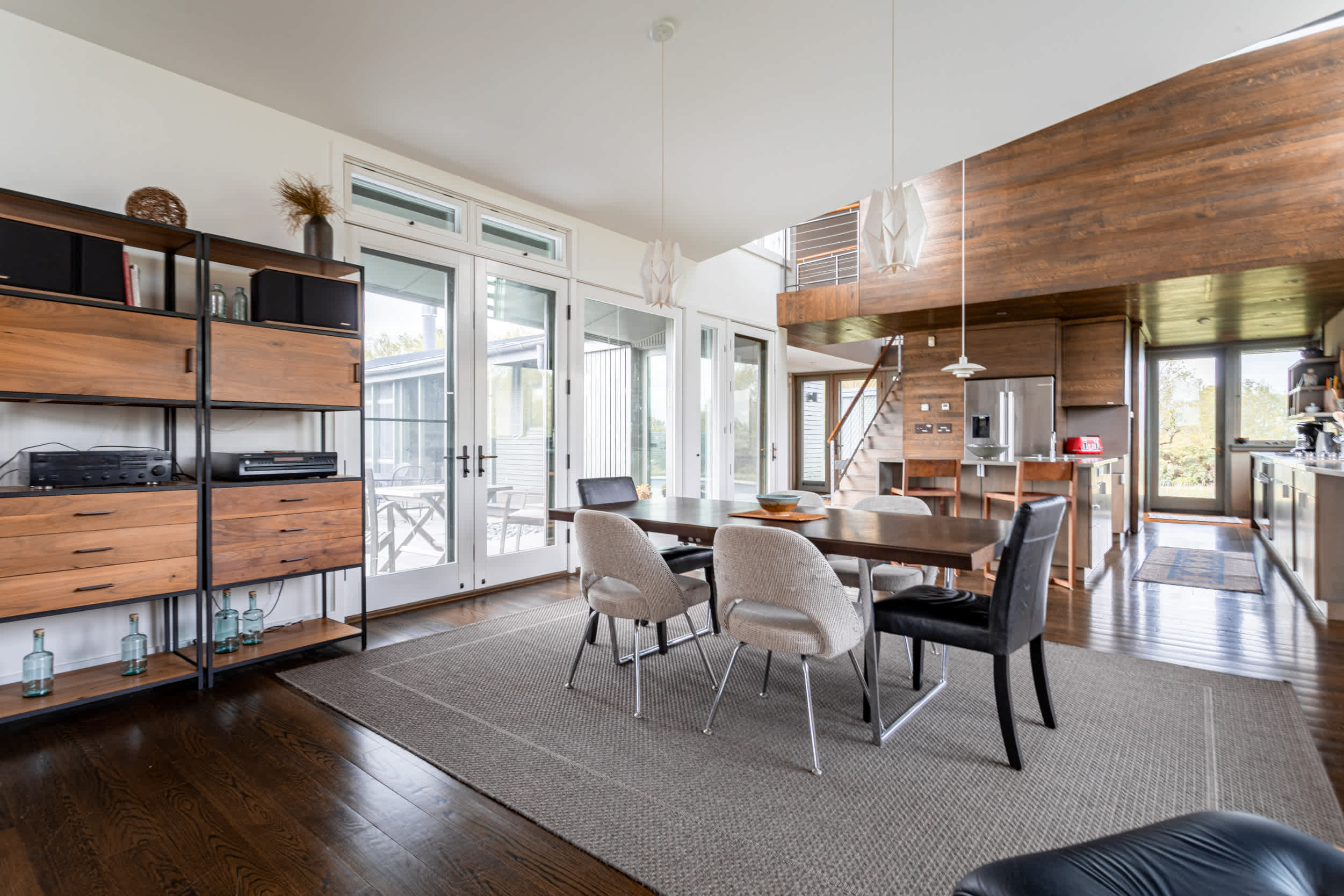 Modern dining area with a dark wood table and mixed chairs on a light grey rug, featuring a tall wooden shelving unit and large glass doors leading to the outdoors.