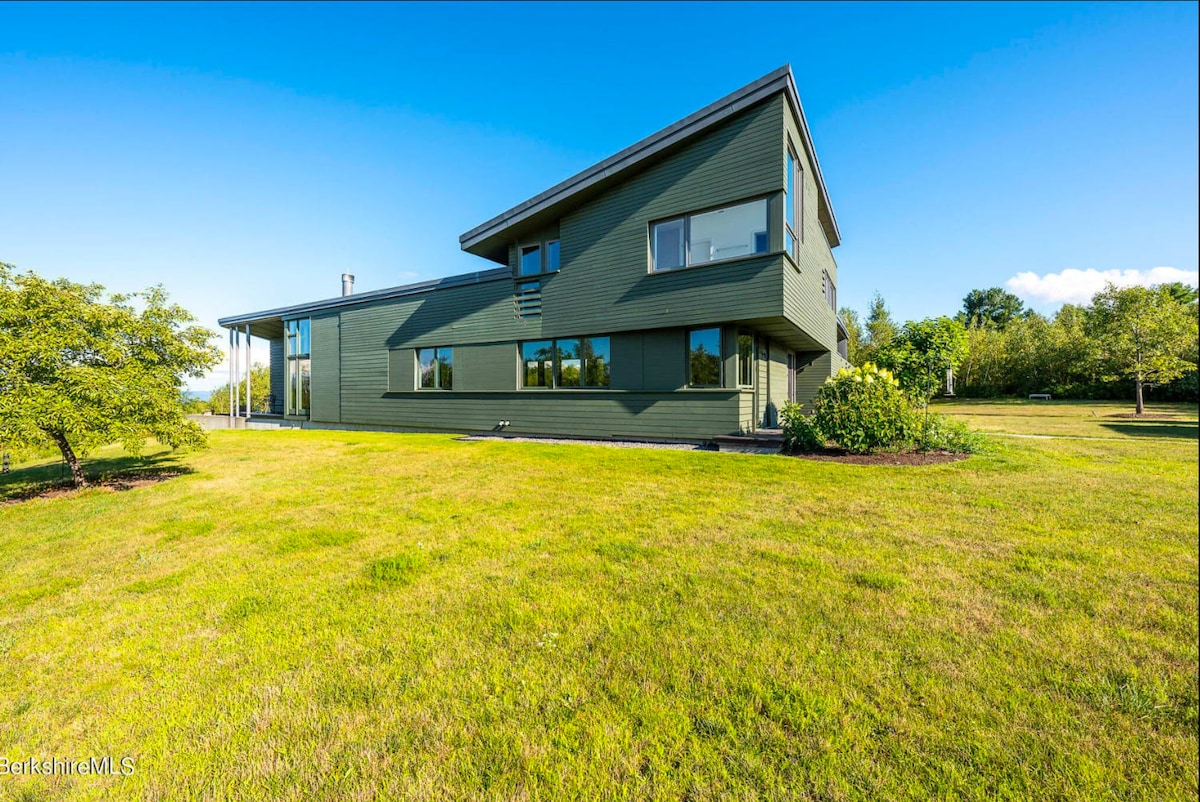 The modern exterior of the house, painted in a muted green, with a distinctive angled roofline and multiple windows. The house is set on a well-maintained green lawn under a clear blue sky.