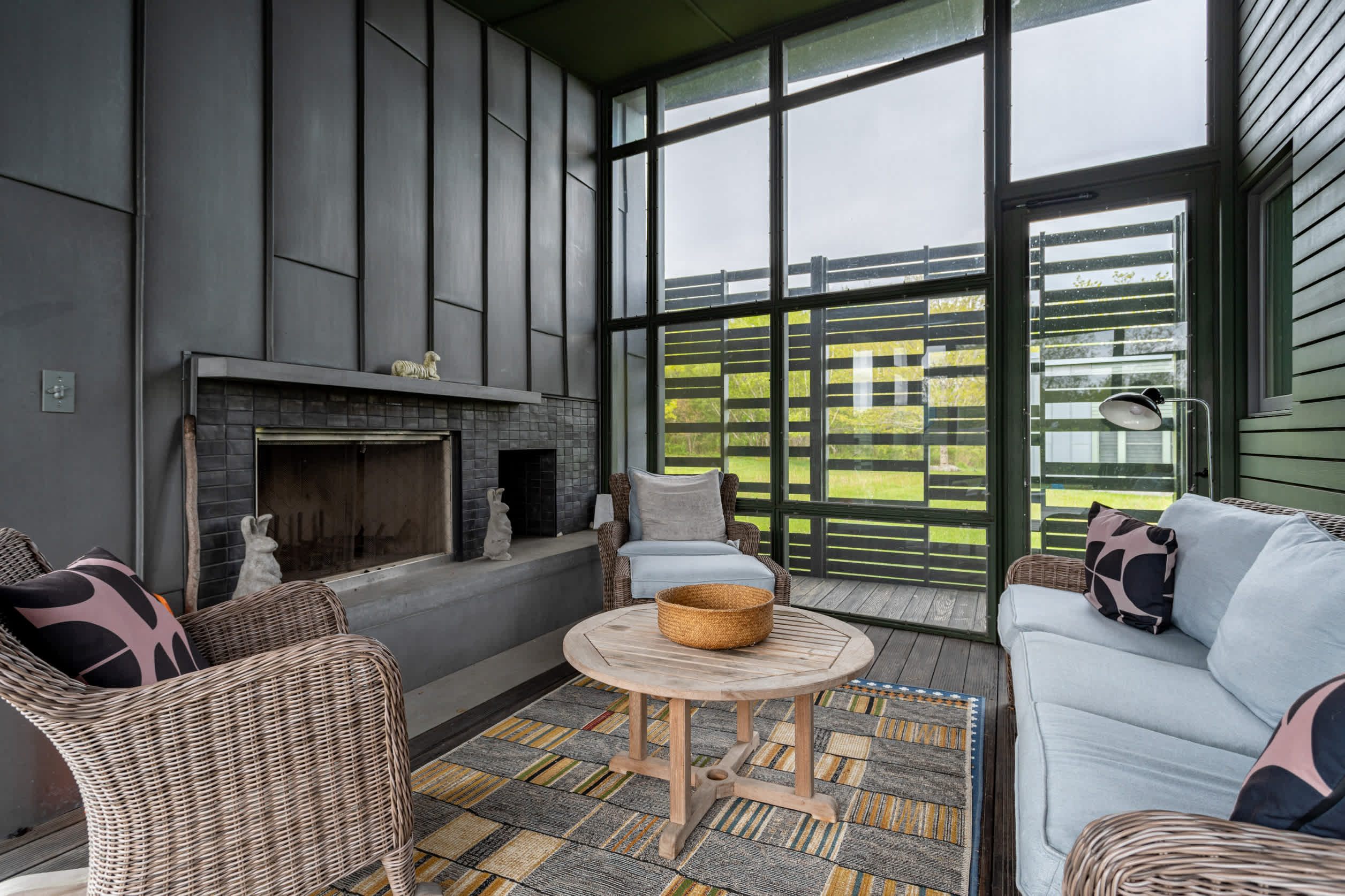 A screened porch features wicker furniture, a patterned rug, and a fireplace with a dark mantel, offering a comfortable outdoor living space with views through the screens to the surrounding greenery.