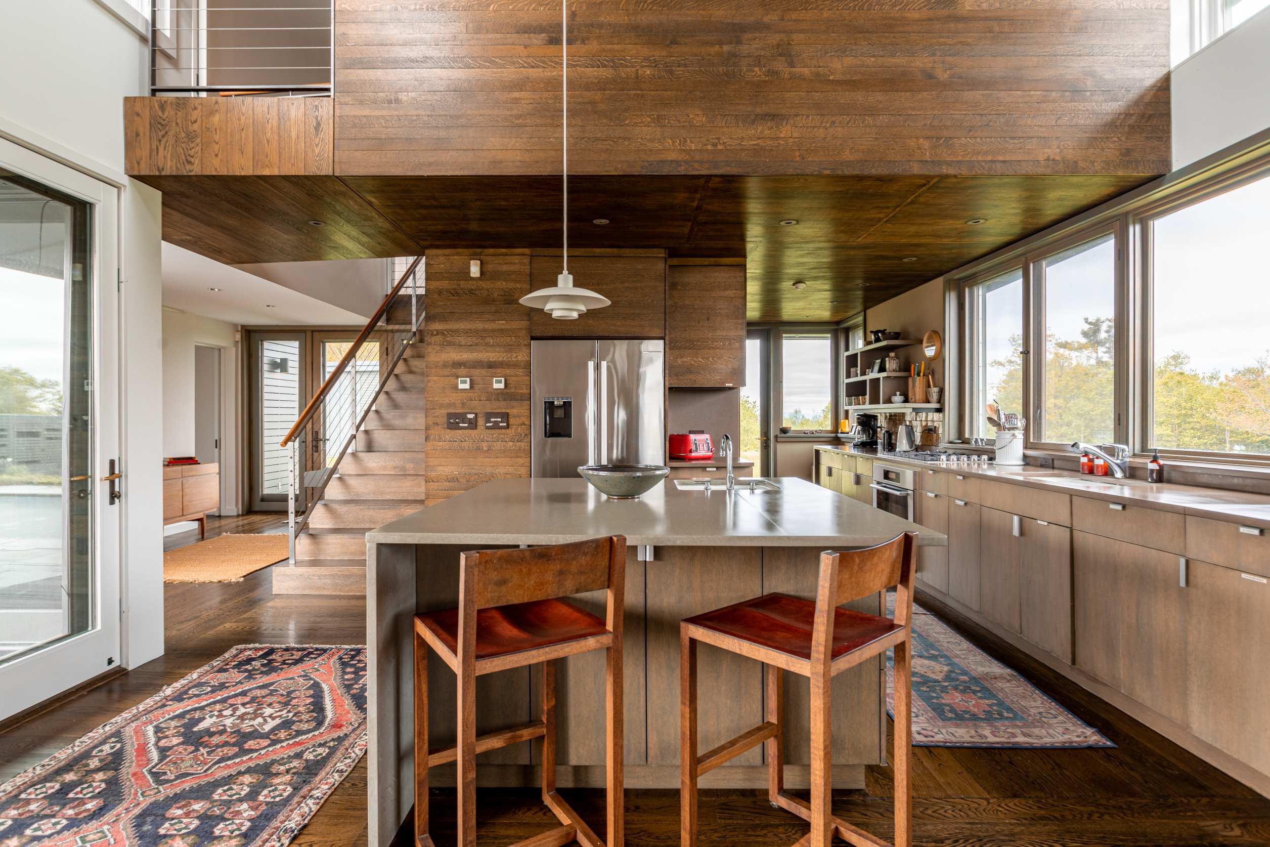 A spacious open-concept kitchen features dark wood floors, light wood cabinetry, a large island with seating, and stainless steel appliances, with a staircase leading to an upper level and large windows offering outdoor views.