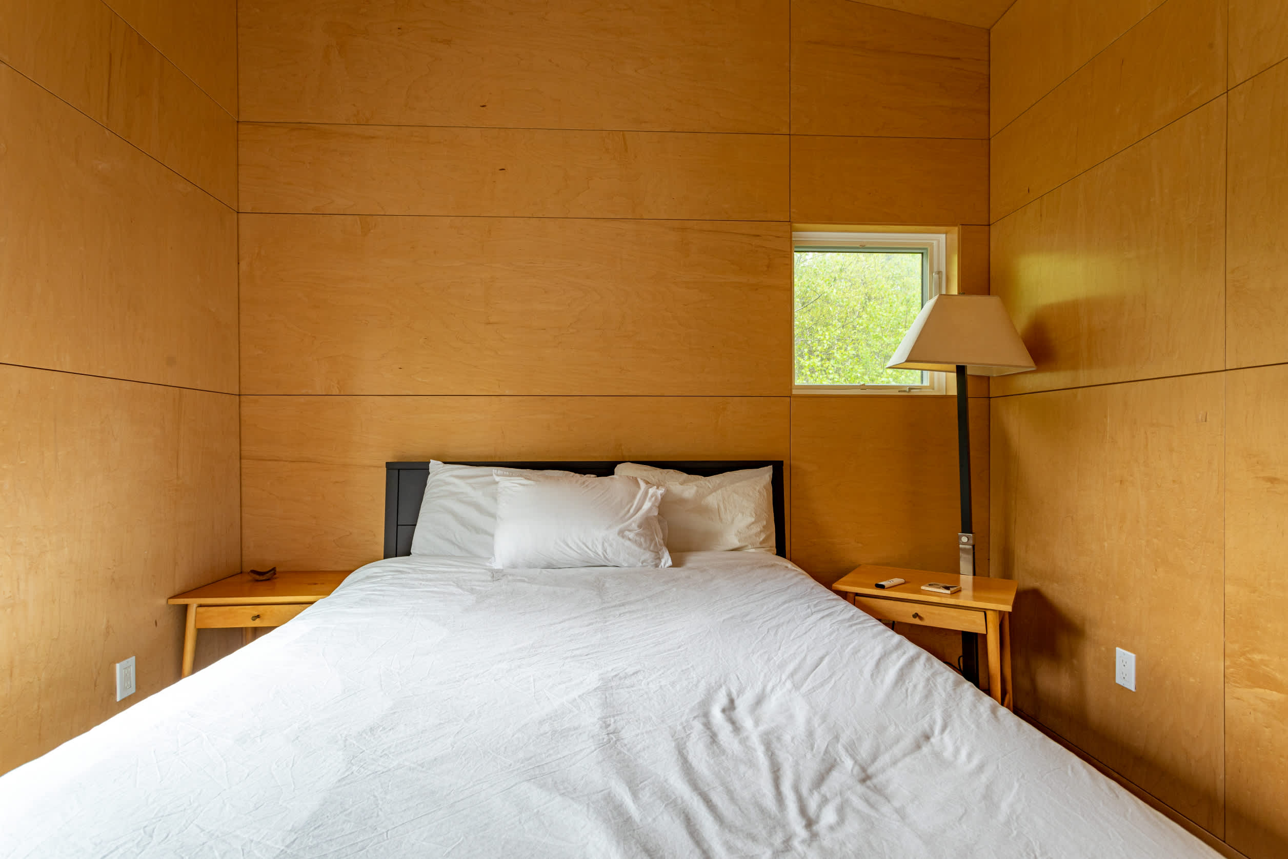 A cozy bedroom with light wood paneling on the walls and ceiling, featuring a bed with white bedding, two wooden nightstands, and a window showing greenery. A tall floor lamp stands next to the bed.