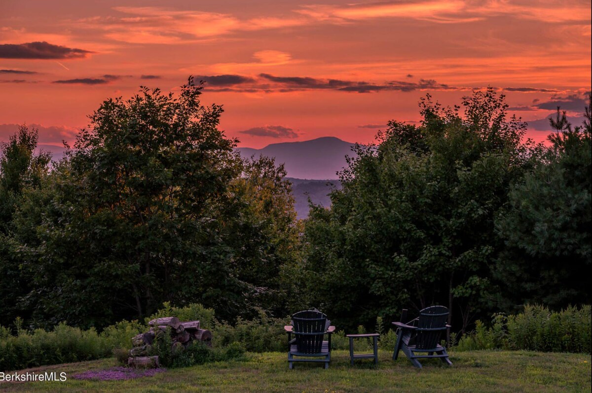 Two dark Adirondack chairs and a small table are positioned on a grassy lawn, facing a breathtaking sunset with vibrant orange and red hues over distant mountains. Lush green trees frame the panoramic view.