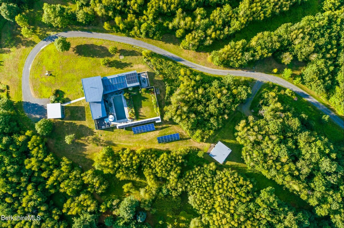 Aerial view of a modern house with a dark roof and solar panels, featuring a rectangular swimming pool and a winding driveway, nestled amidst lush green forest. The property also includes a separate small shed and additional ground-mounted solar panels.