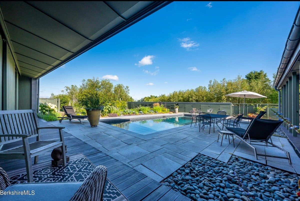 An expansive outdoor patio area with a rectangular swimming pool, surrounded by dark grey pavers and decking. Various lounge chairs, an outdoor dining set, and lush garden beds are visible under a bright blue sky.