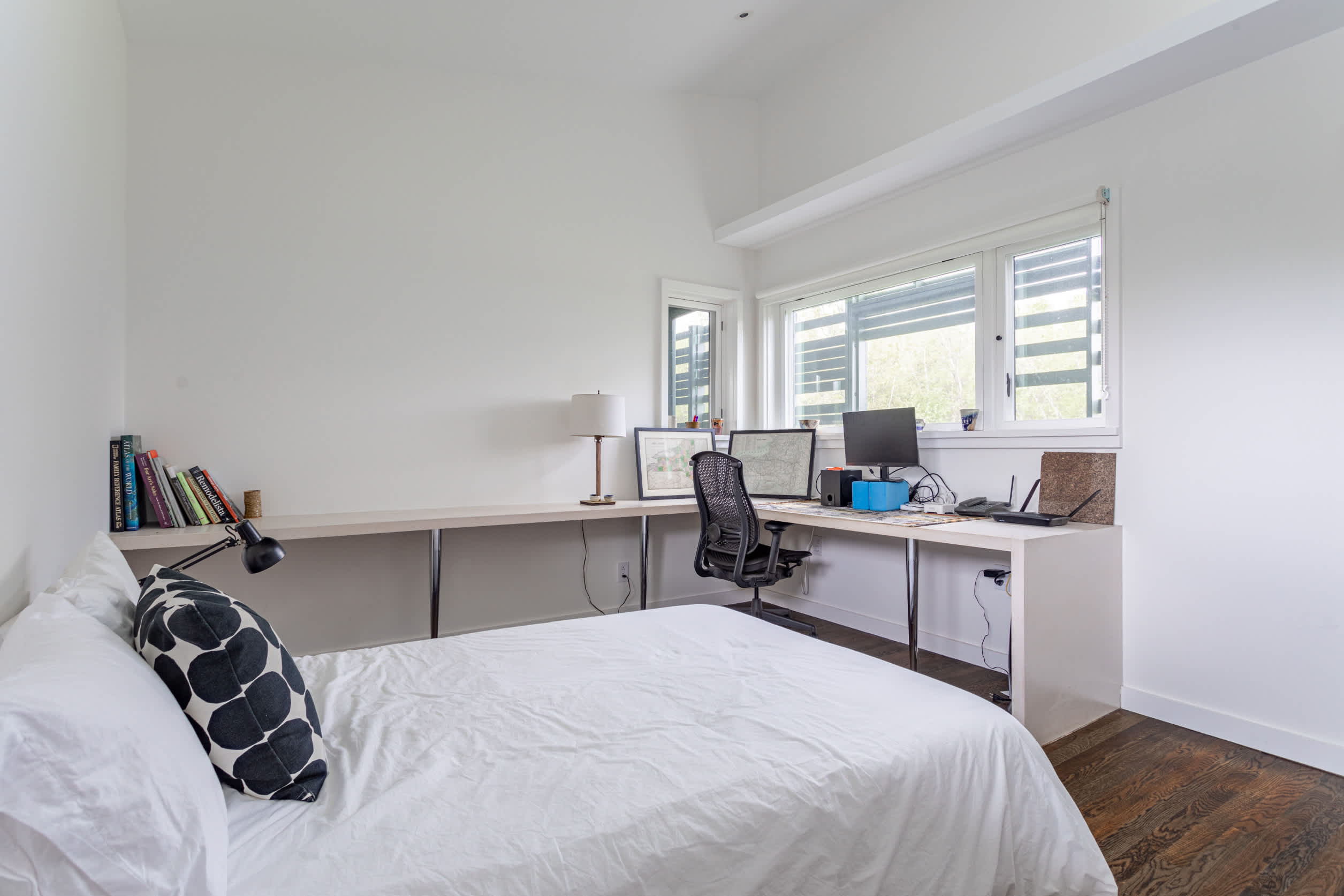 Modern bedroom featuring a white bed with a black and white patterned pillow, dark wood floors, and a long built-in desk with two computer monitors. Large windows provide natural light, and a black office chair is present.